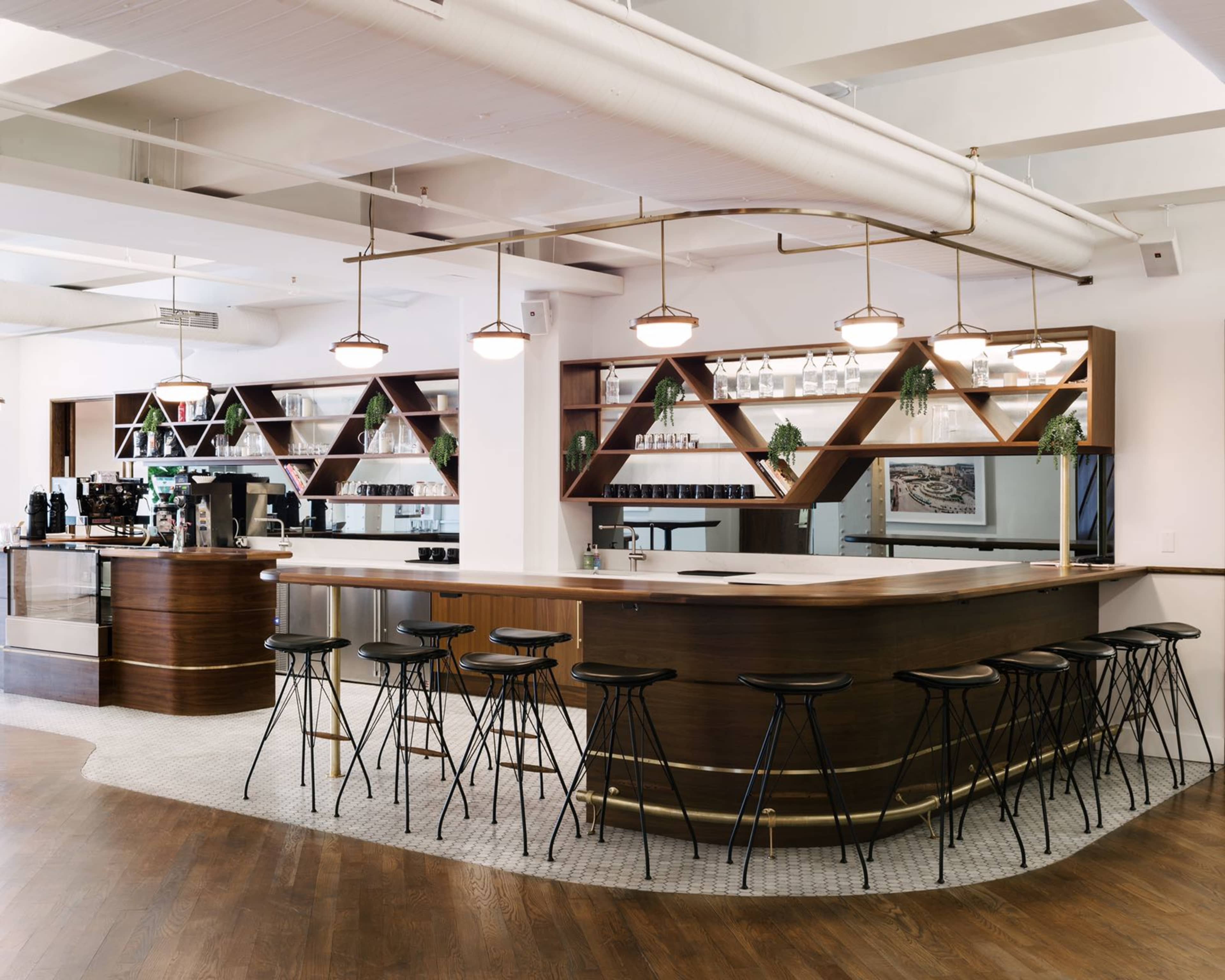 A modern bar area with a wooden counter, black stools, and geometric shelves displaying glassware and plants.