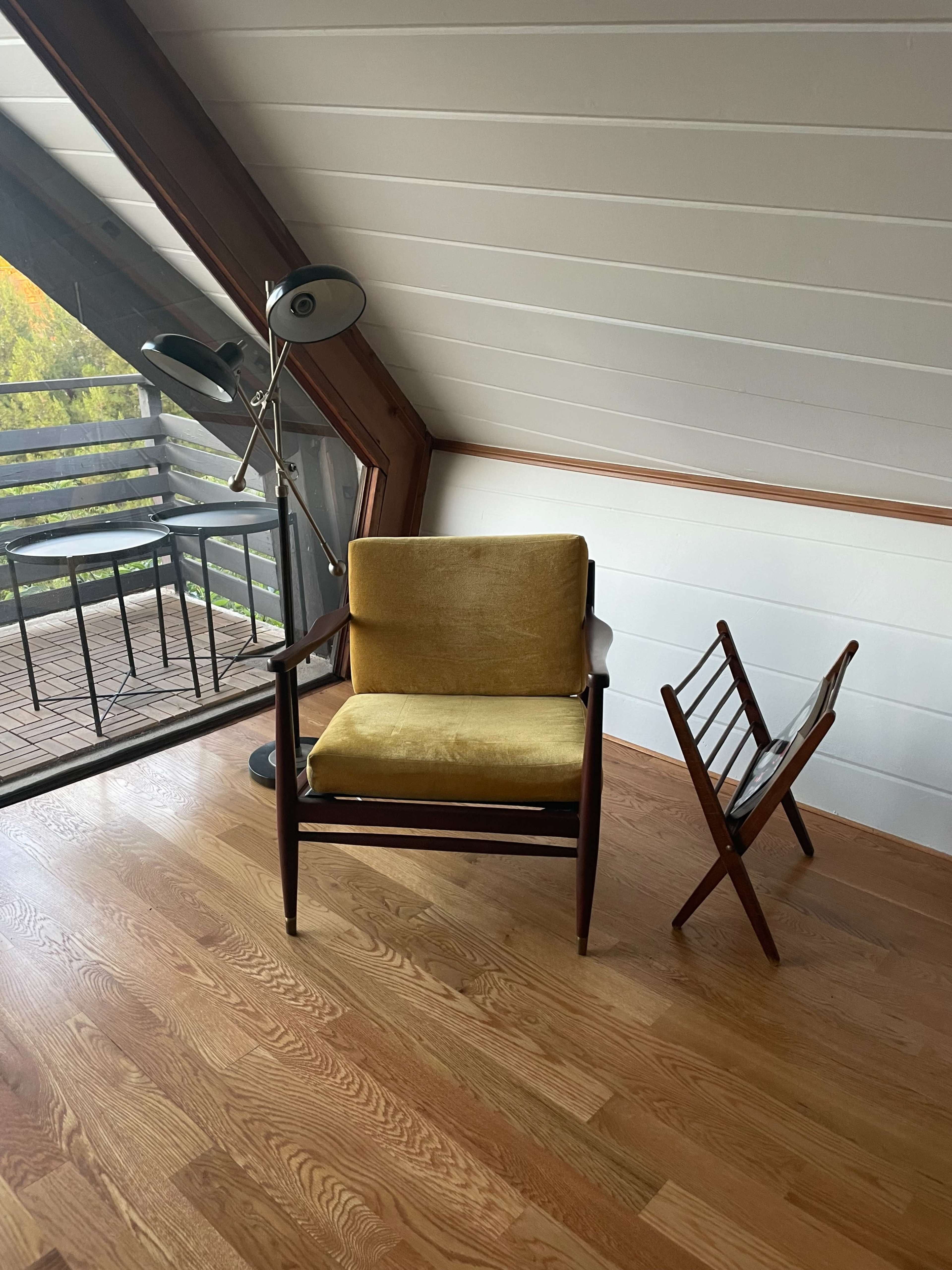 A yellow upholstered chair and a folding wooden chair are positioned in a corner of a room with sloped wooden ceilings and a balcony visible through a large window.