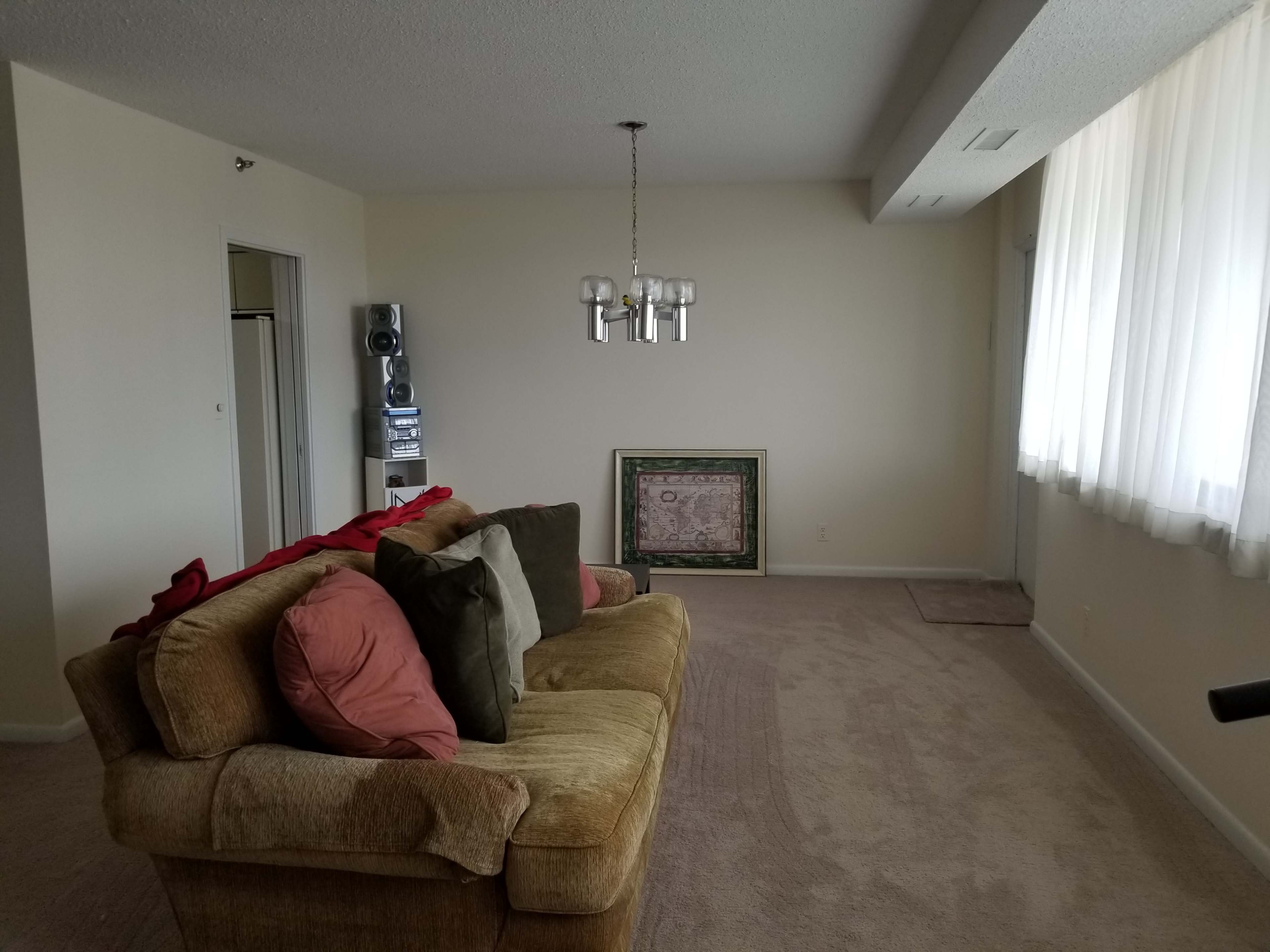 A sparsely furnished living room with a brown sofa featuring colorful cushions, a pendant light, and a framed artwork leaning against the wall.