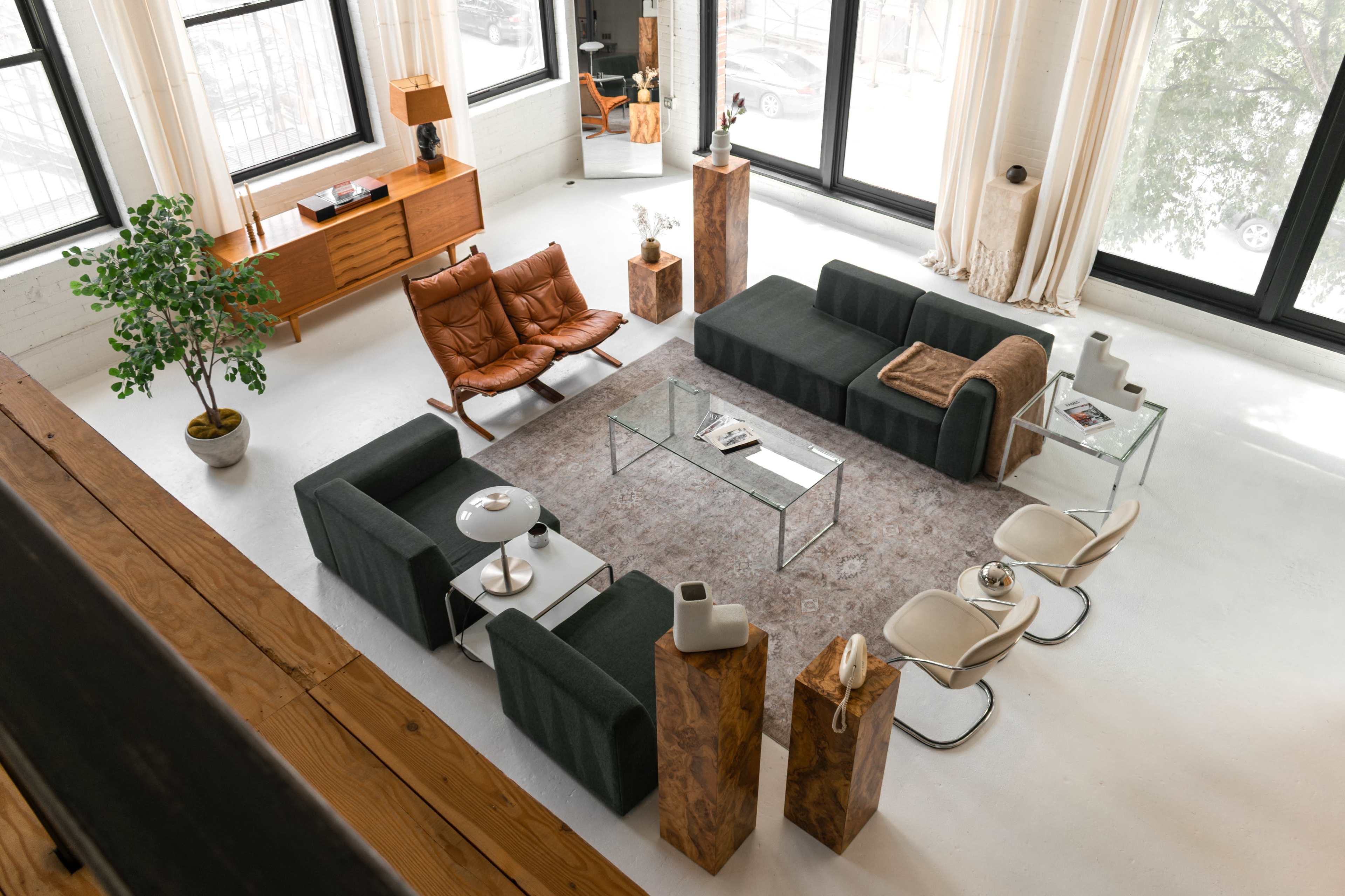 The image shows a spacious living room featuring modern furniture, including a glass coffee table, two dark green sofas, a brown lounge chair, and two white chairs, all arranged around a large area rug.