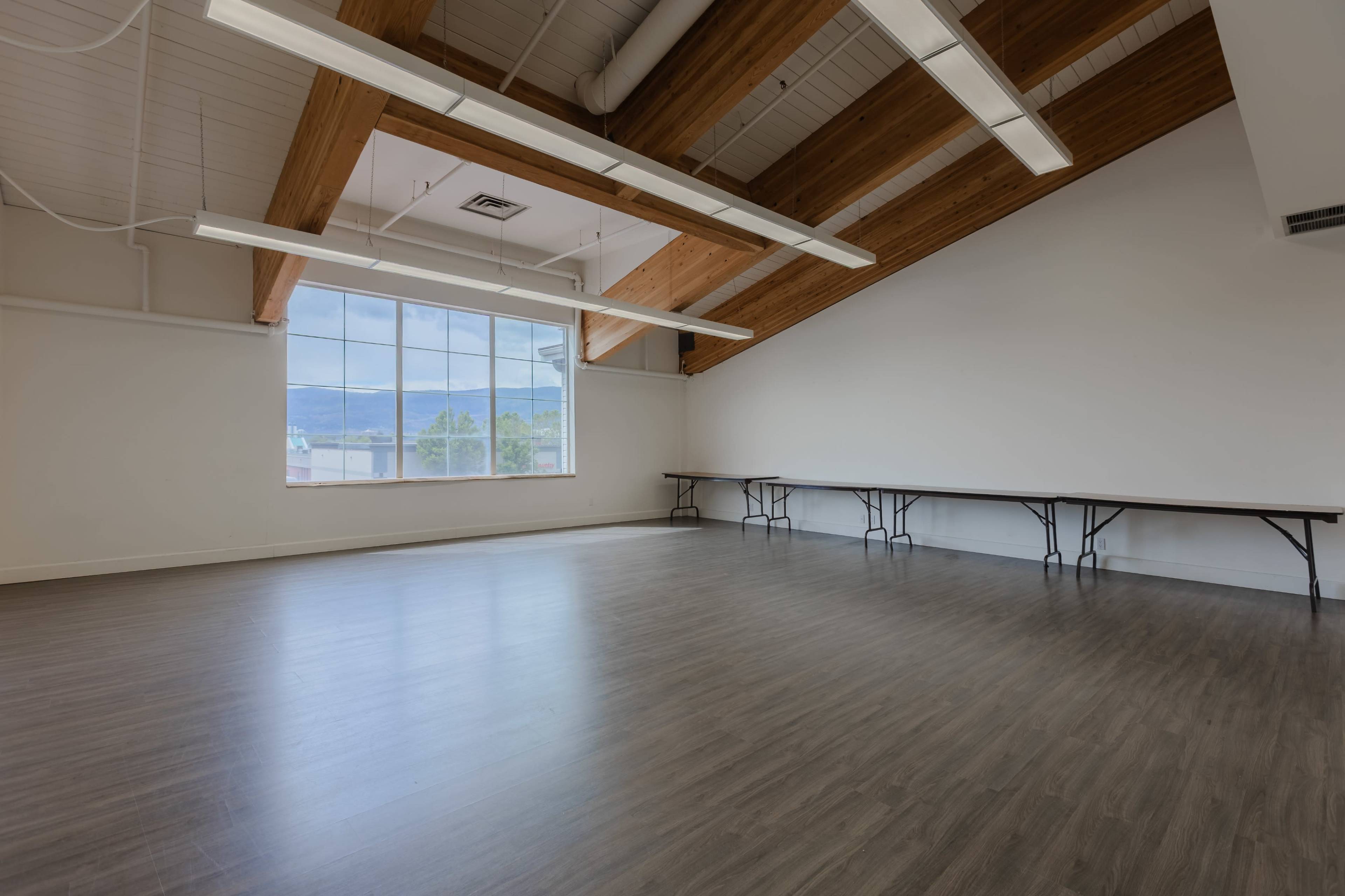A spacious, empty room with a high ceiling and large windows, featuring wooden beams and tables along one wall.