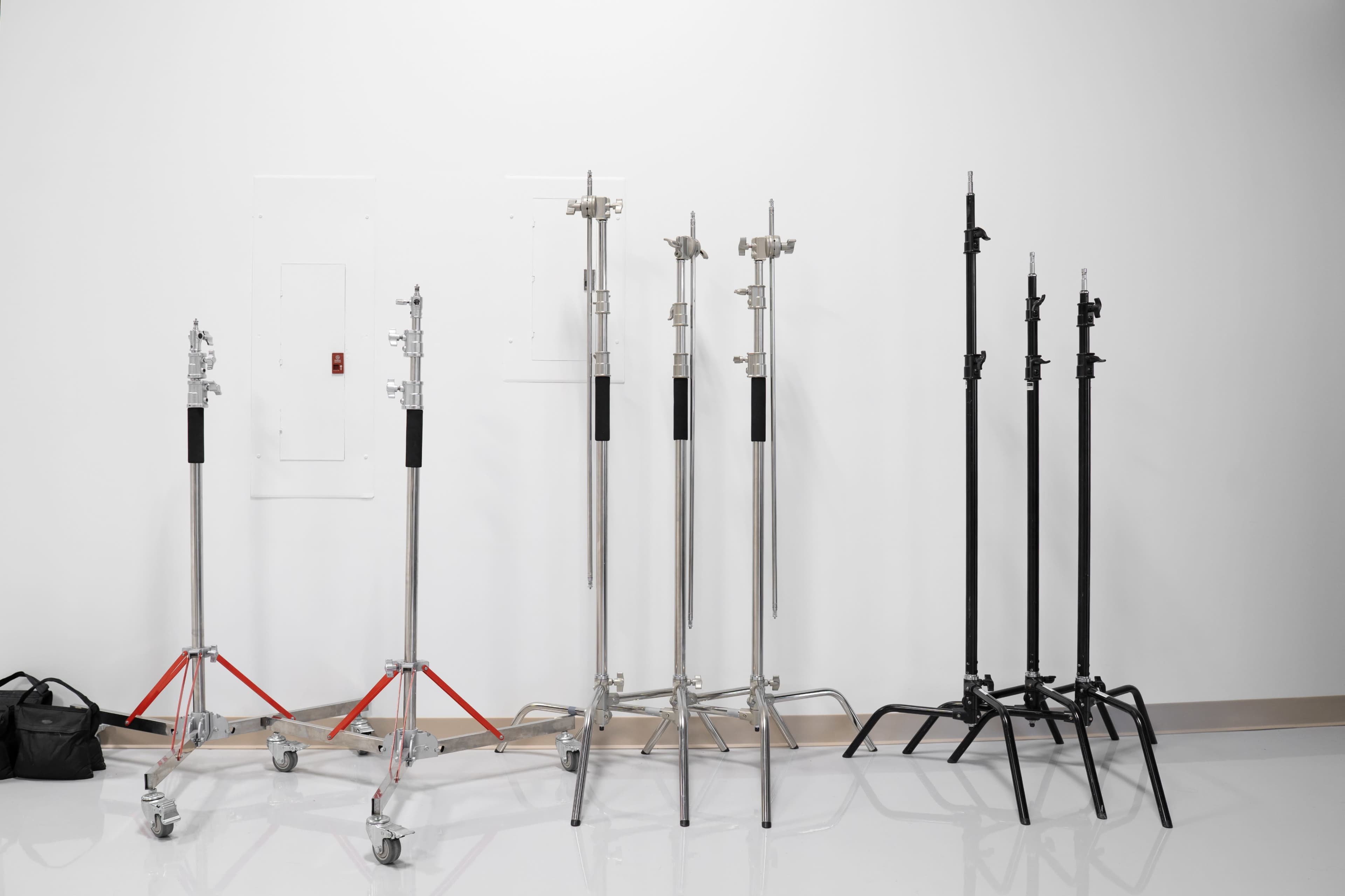 The image shows a lineup of various photography light stands, including silver and black models, arranged against a plain white wall.