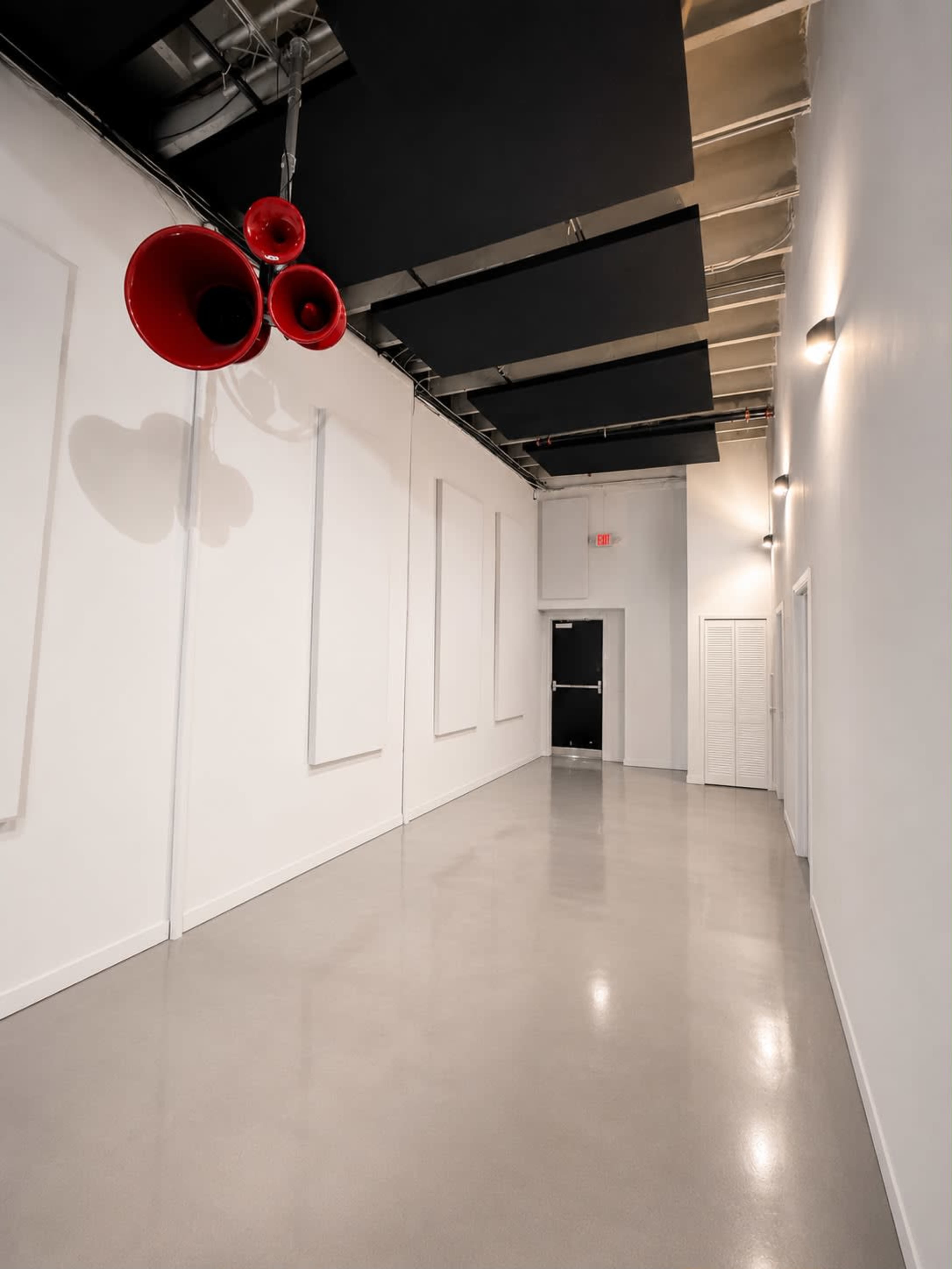 The image shows a long, narrow hallway with a polished concrete floor, white walls, and three red speakers hanging from the ceiling.