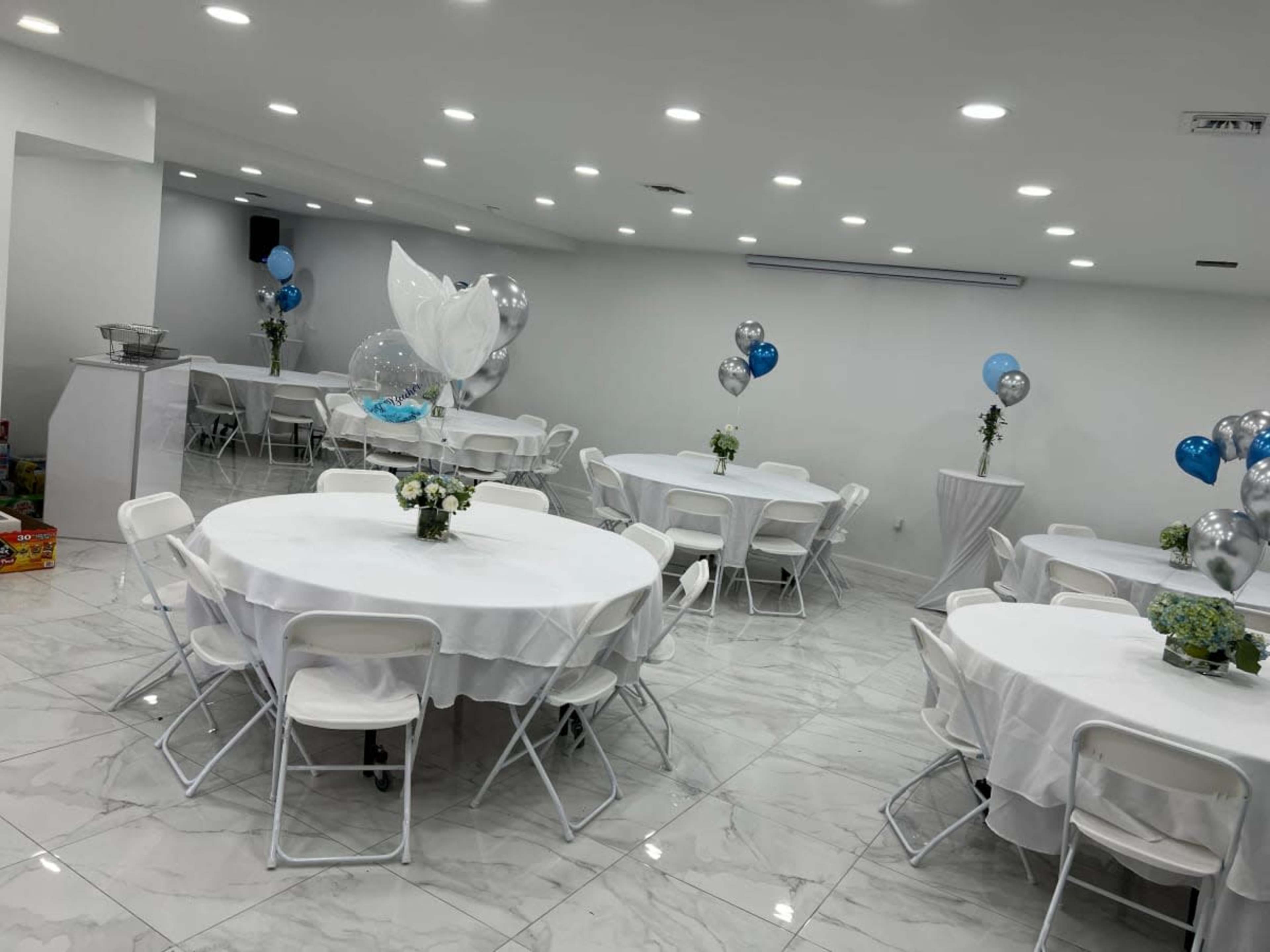 The image shows a brightly lit event space with several round tables covered in white tablecloths, decorated with centerpieces and surrounded by balloons.