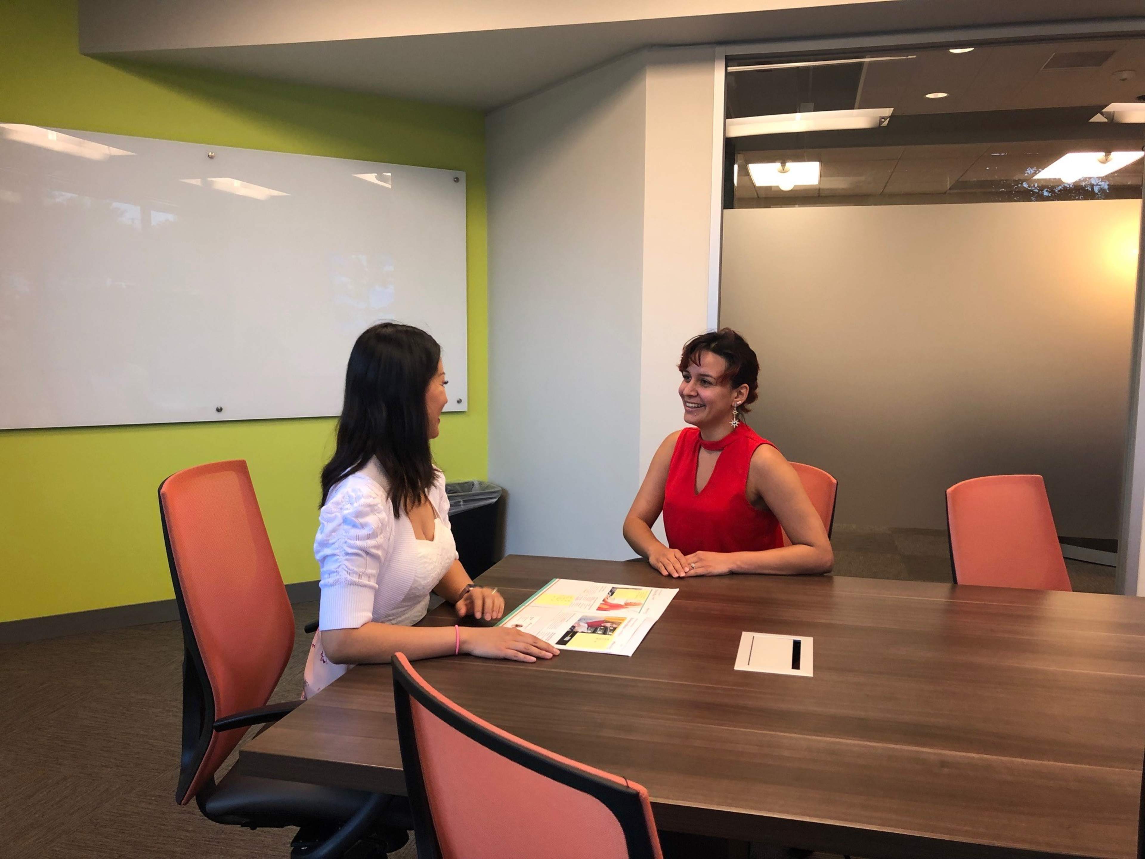 Two women are seated at a wooden conference table in a modern meeting room, discussing documents.