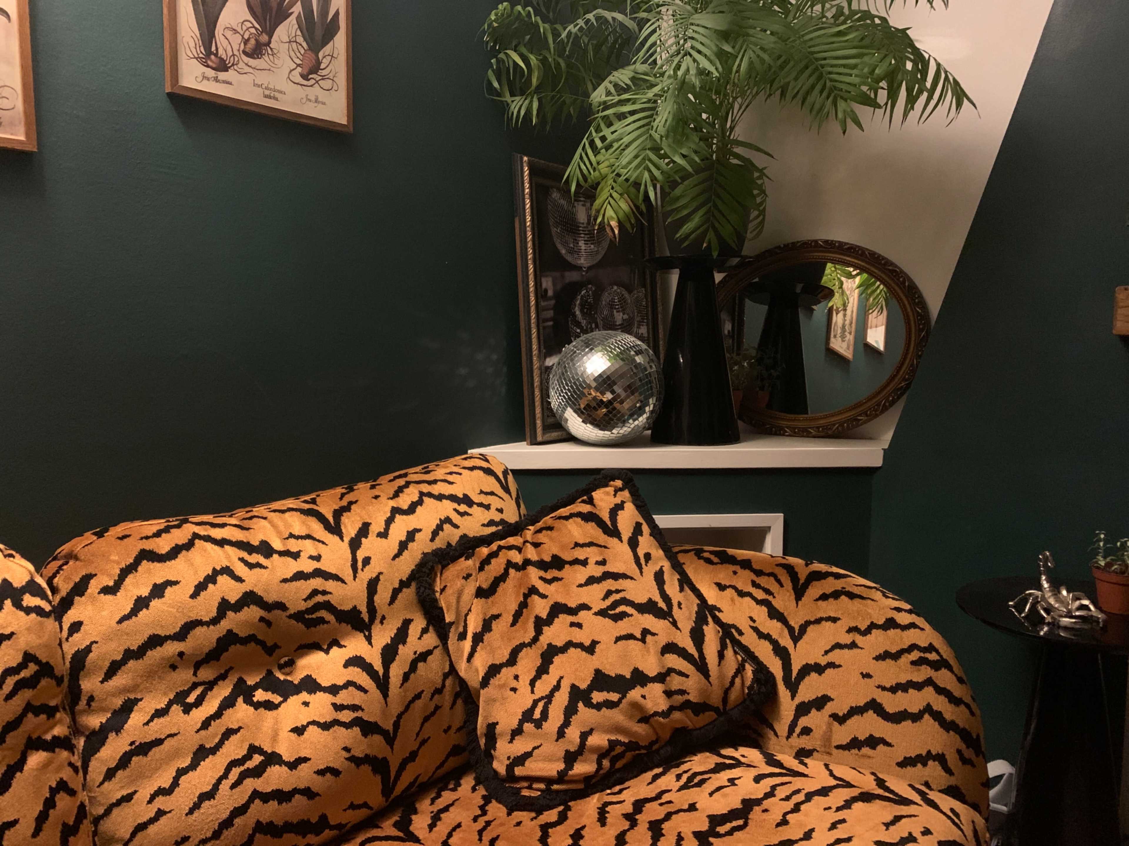 The image shows a corner of a room featuring a tiger-striped couch, a potted plant, a disco ball, and framed artwork on a dark green wall.