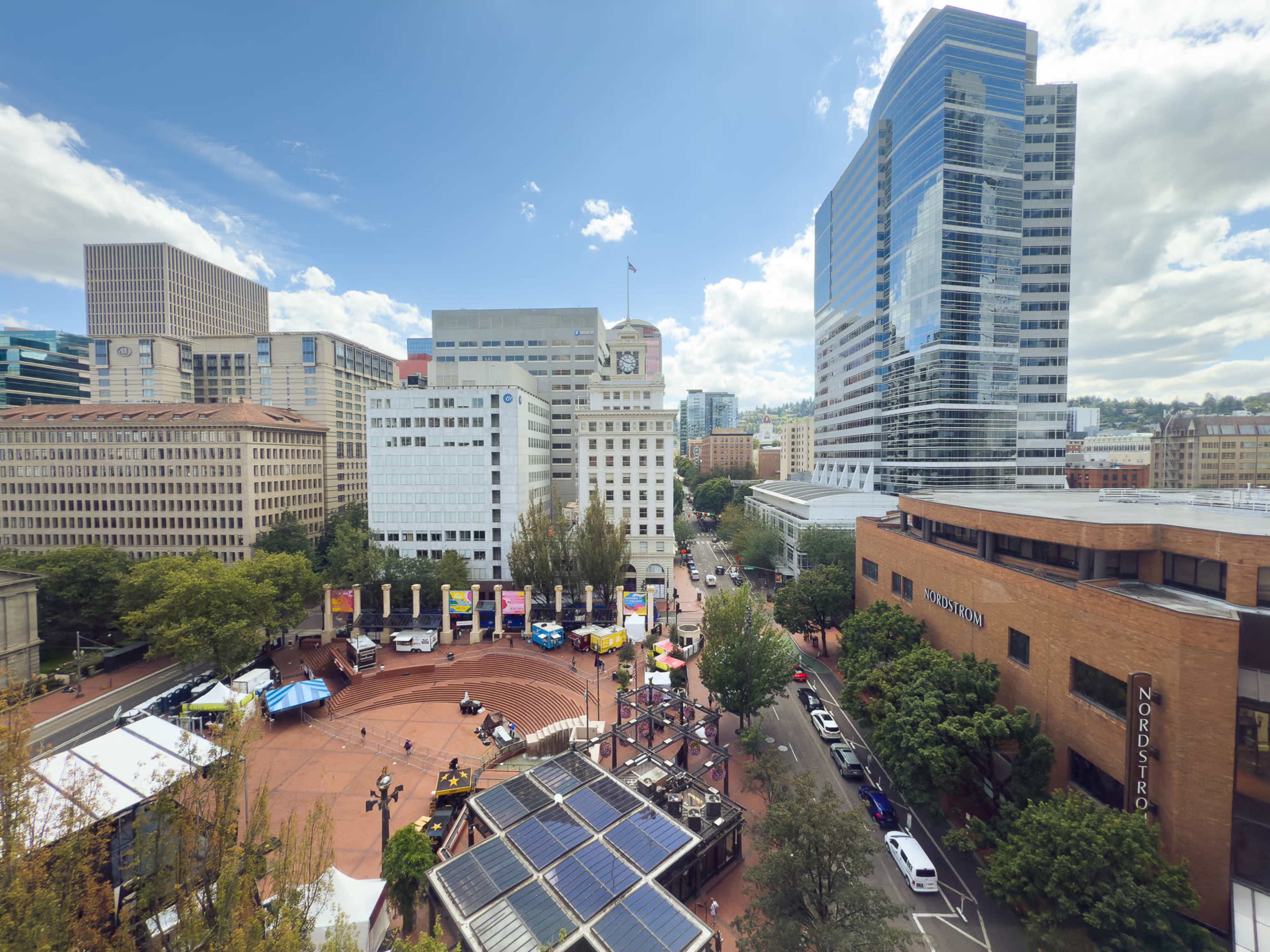 The image shows a bustling urban plaza with buildings, trees, and various vendors set up, including tents and food stalls.