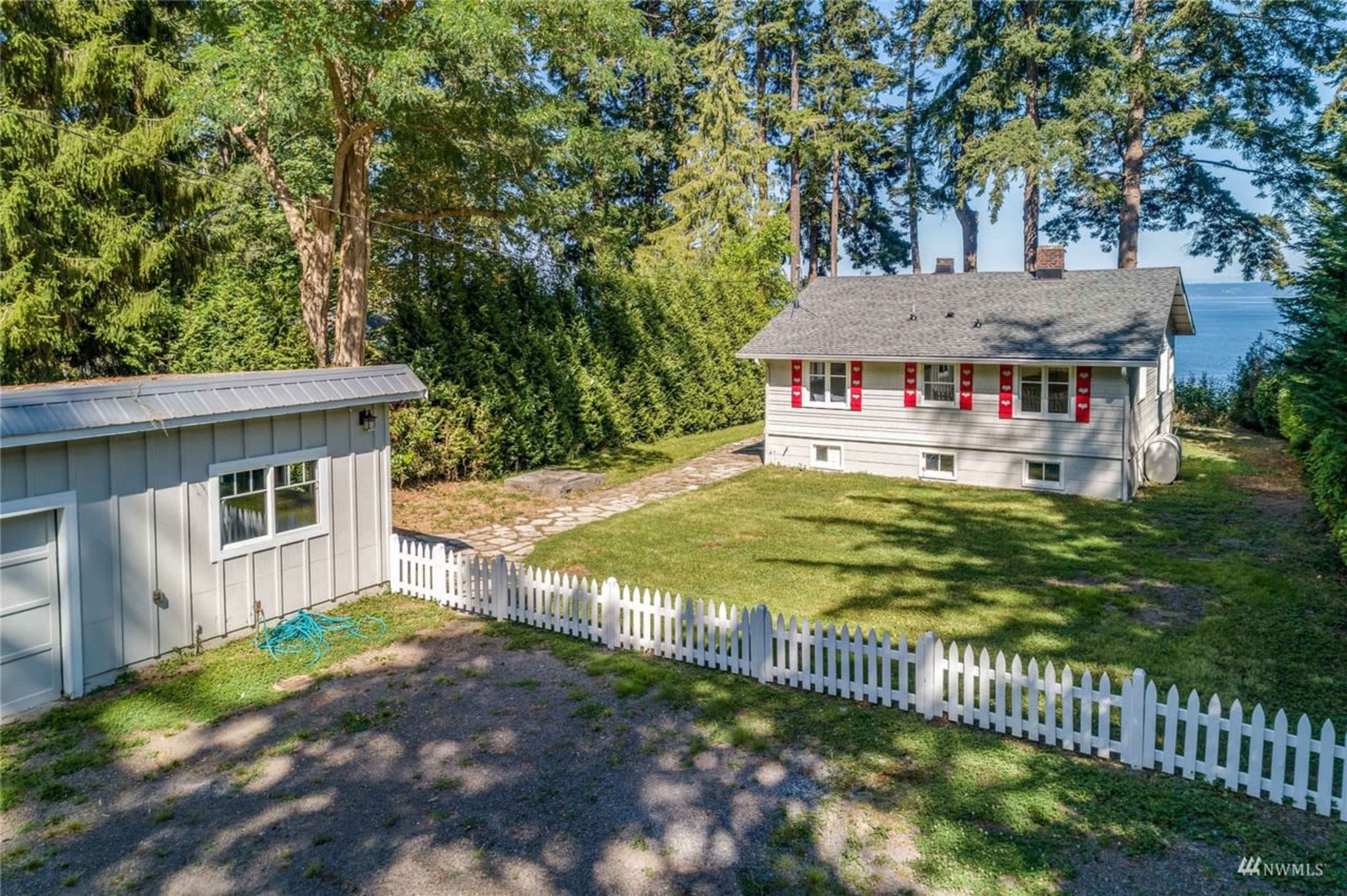 A single-story house with red shutters, surrounded by a green lawn and a white picket fence, with a garage and a stone patio nearby, set against a backdrop of tall trees and a body of water in the distance.