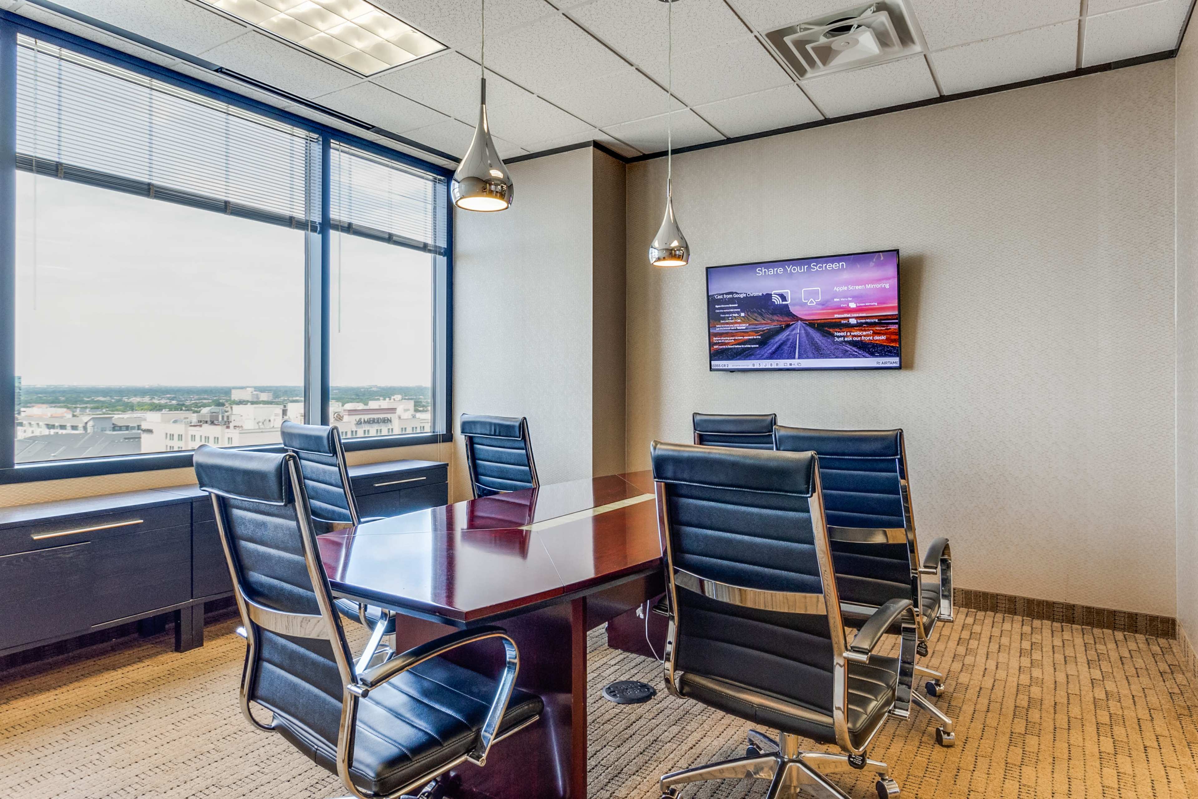 A conference room features a wooden table surrounded by black chairs, with a large window displaying a city view and a mounted screen displaying a digital presentation.