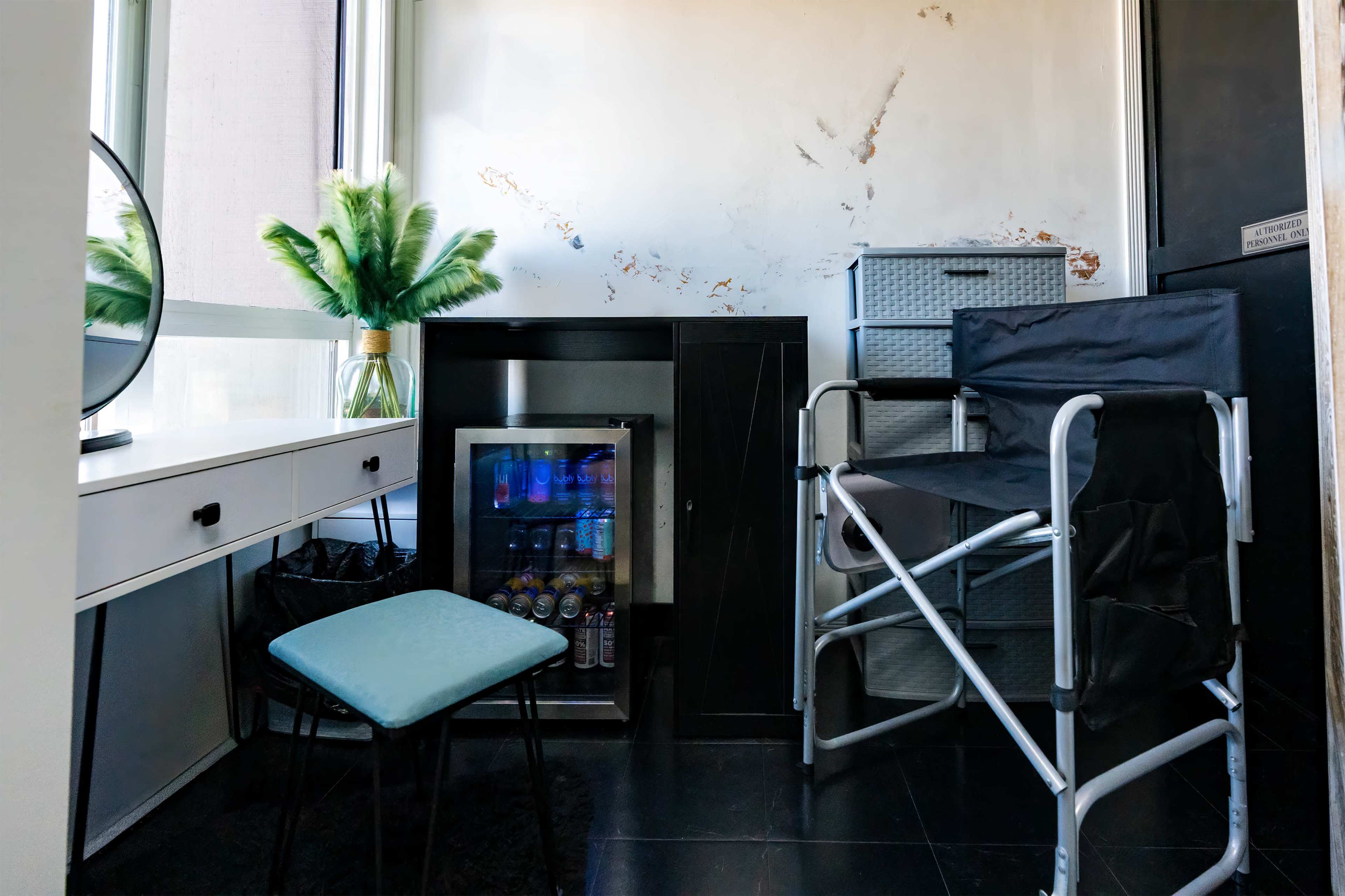 The image shows a small room featuring a desk with a mirror, a mini-fridge, a foldable chair, and a plant in a vase, all against a wall with peeling paint.