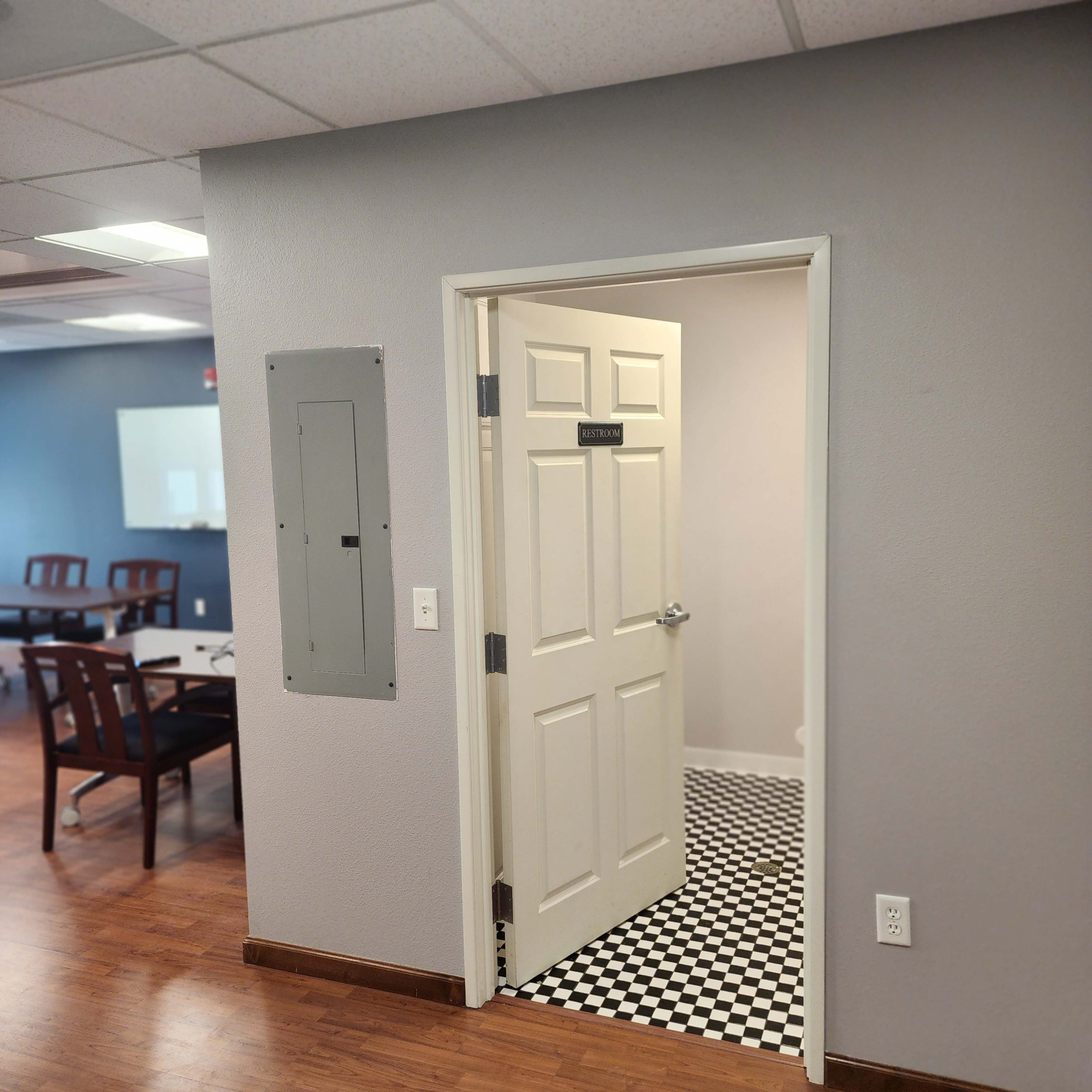 An open door leading to a checkered floor room from a corridor with wooden flooring and a switch panel on the wall.