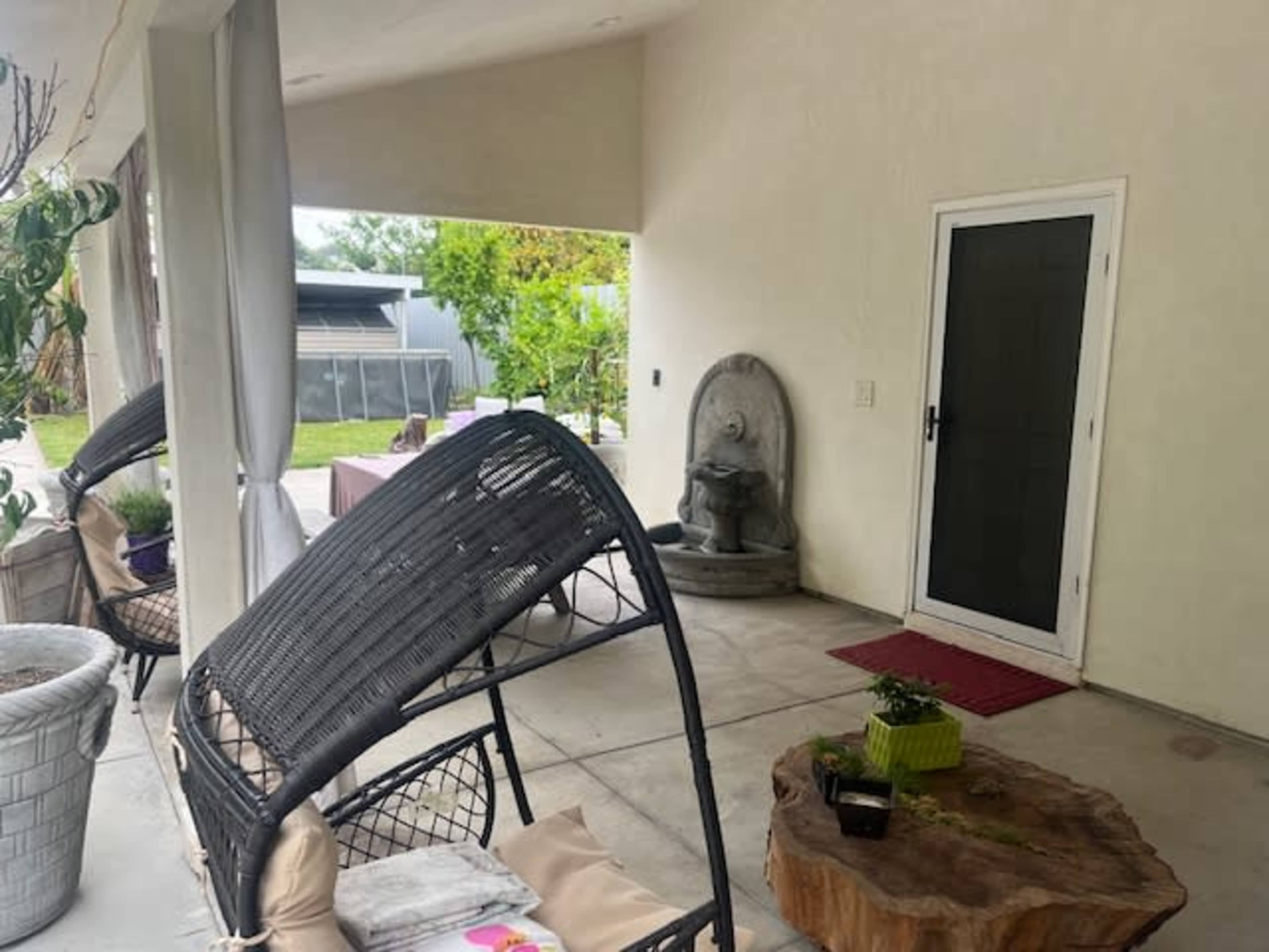 A spacious outdoor patio area with a wicker chair, a wooden side table, and a stone water feature next to a door.
