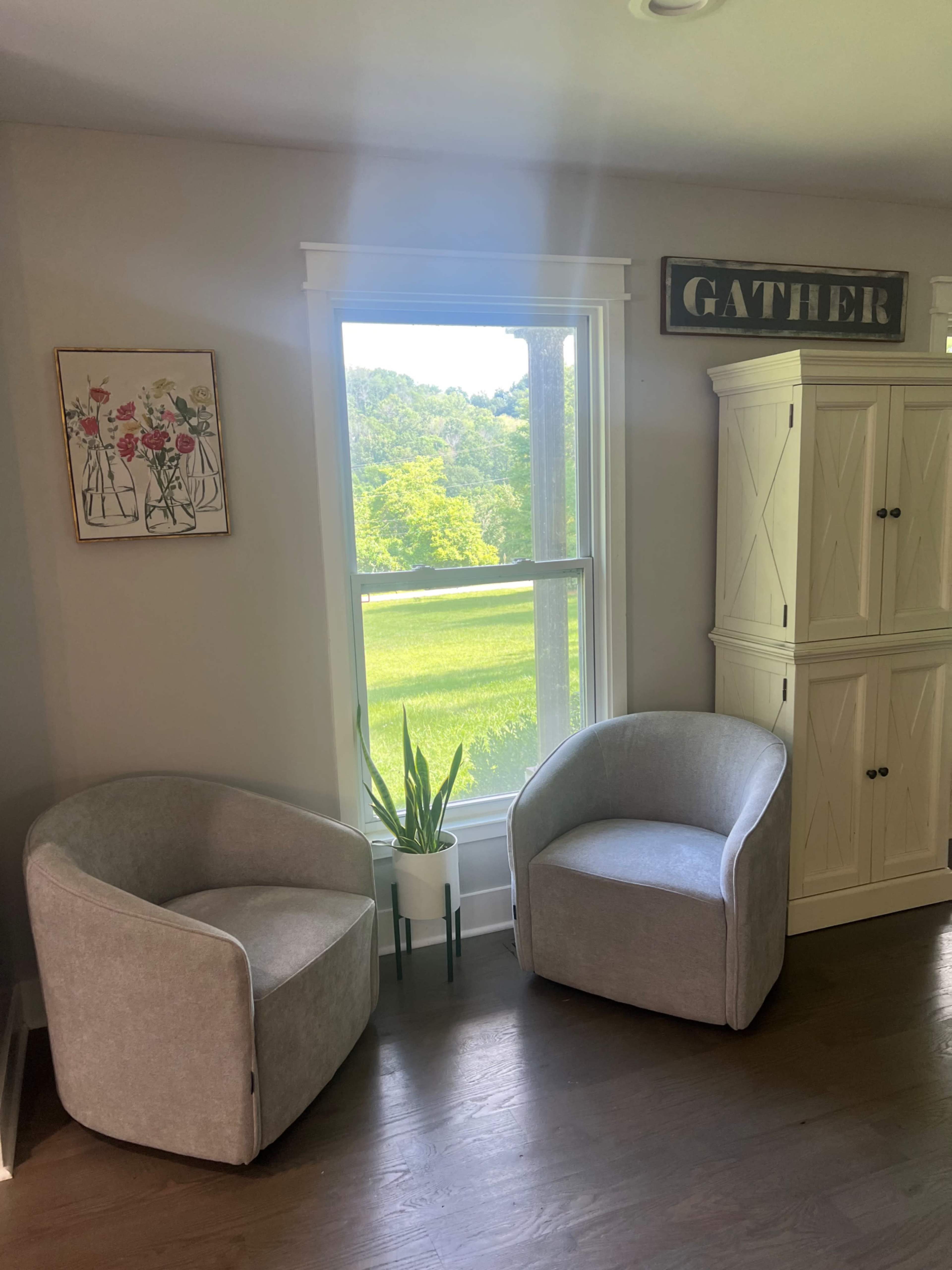 Two upholstered chairs are positioned next to a window with a view of greenery, accompanied by a potted plant and a decorative sign on the wall.