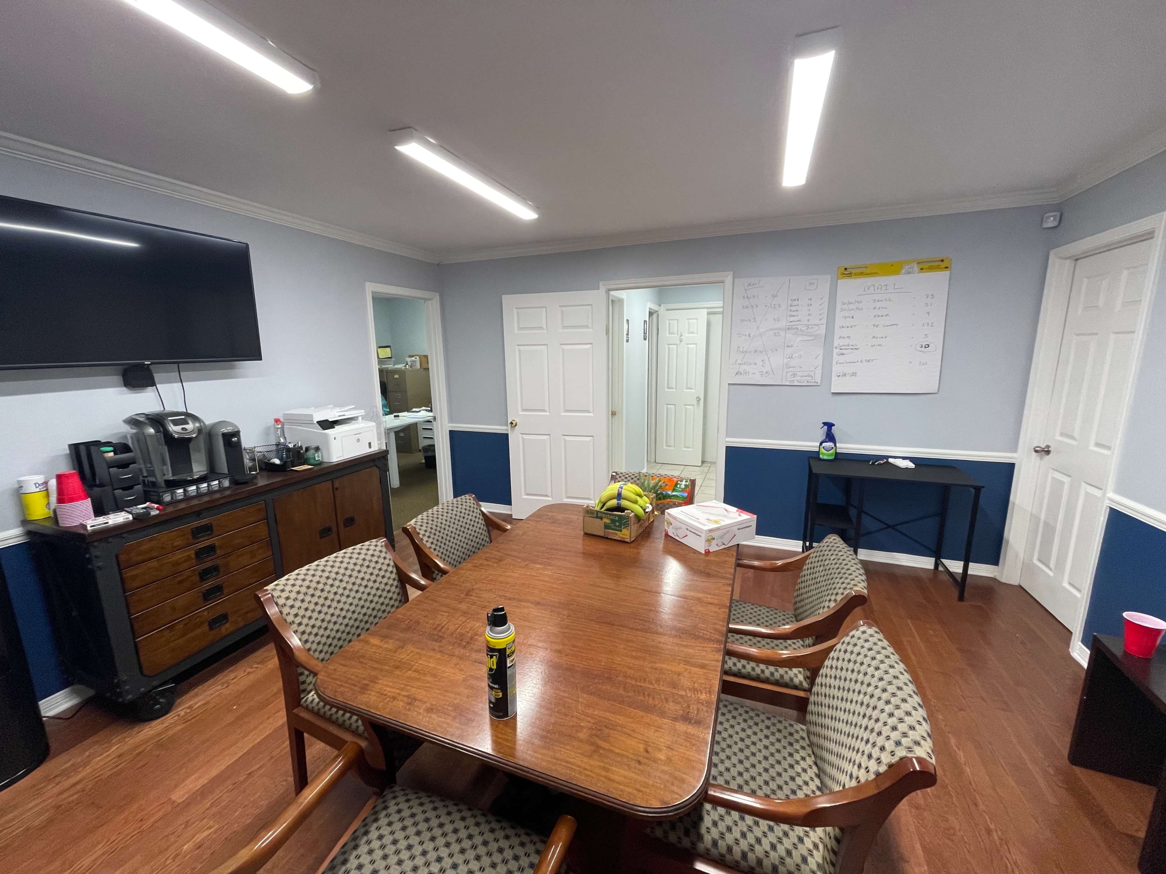 A conference room features a wooden table surrounded by chairs, a television mounted on the wall, and a coffee station with supplies nearby.