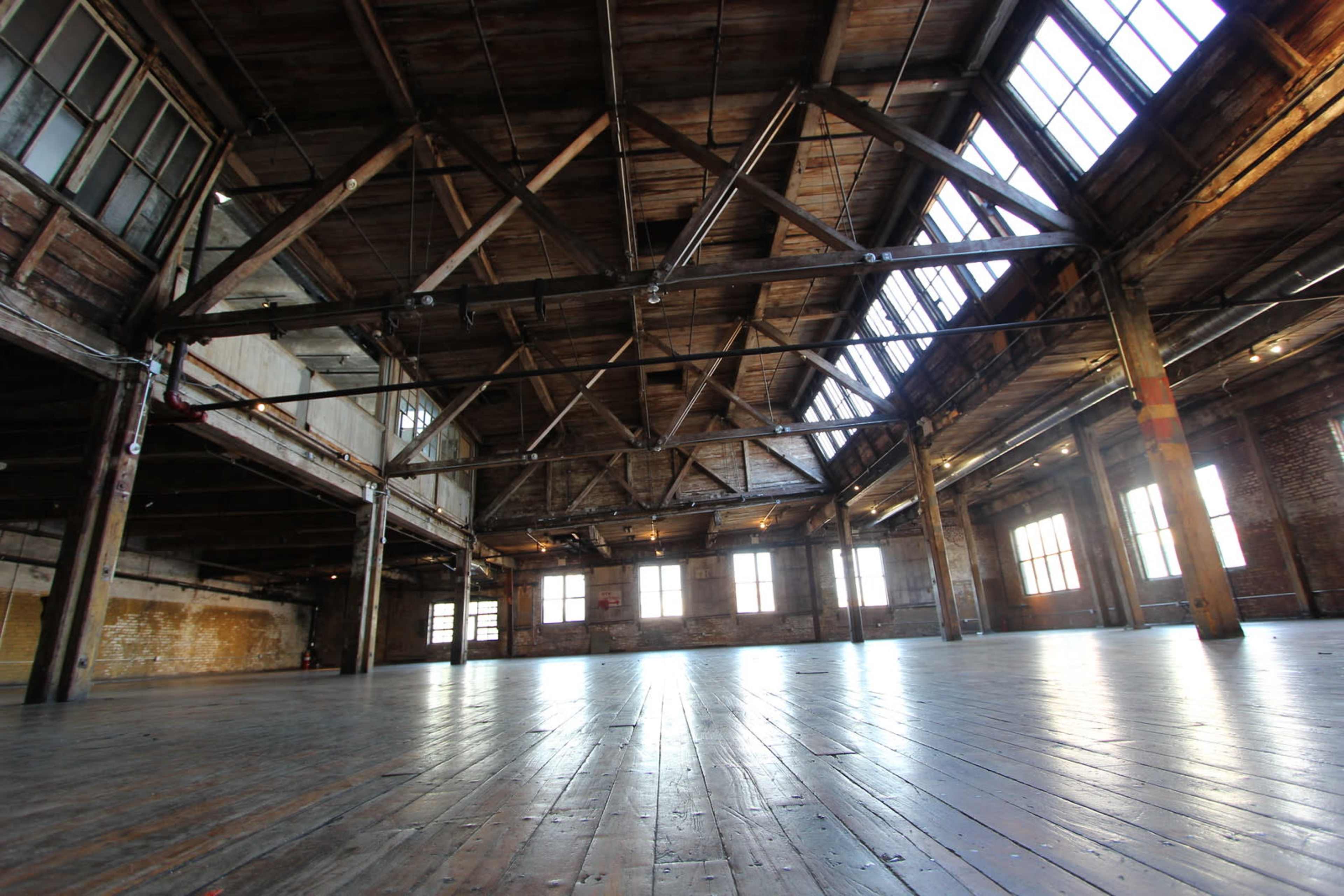 A spacious, industrial interior with wooden beams and large windows illuminating a wooden floor.