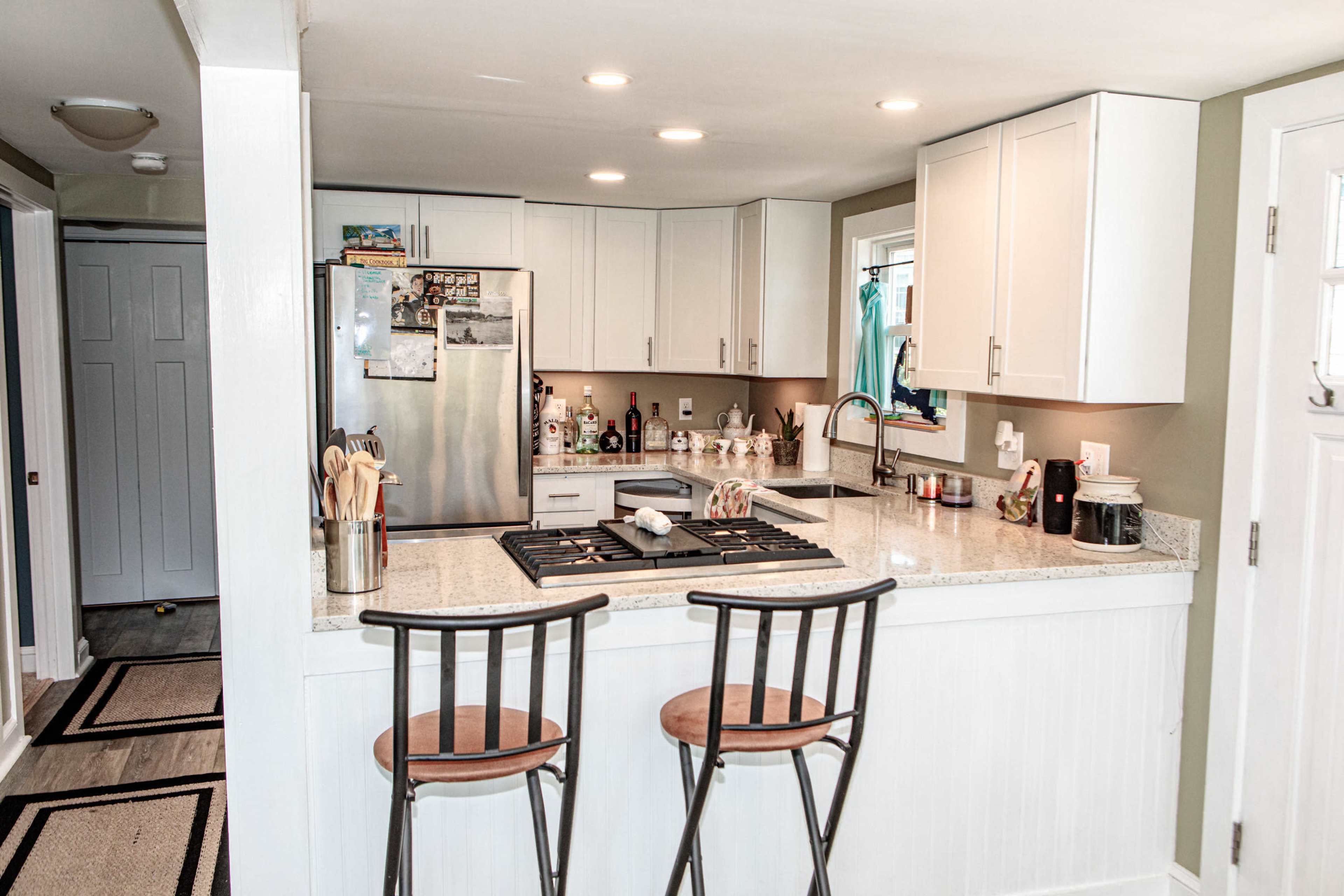 The image shows a modern kitchen featuring white cabinets, a stainless steel refrigerator, a granite countertop with bar stools, and a gas stove.