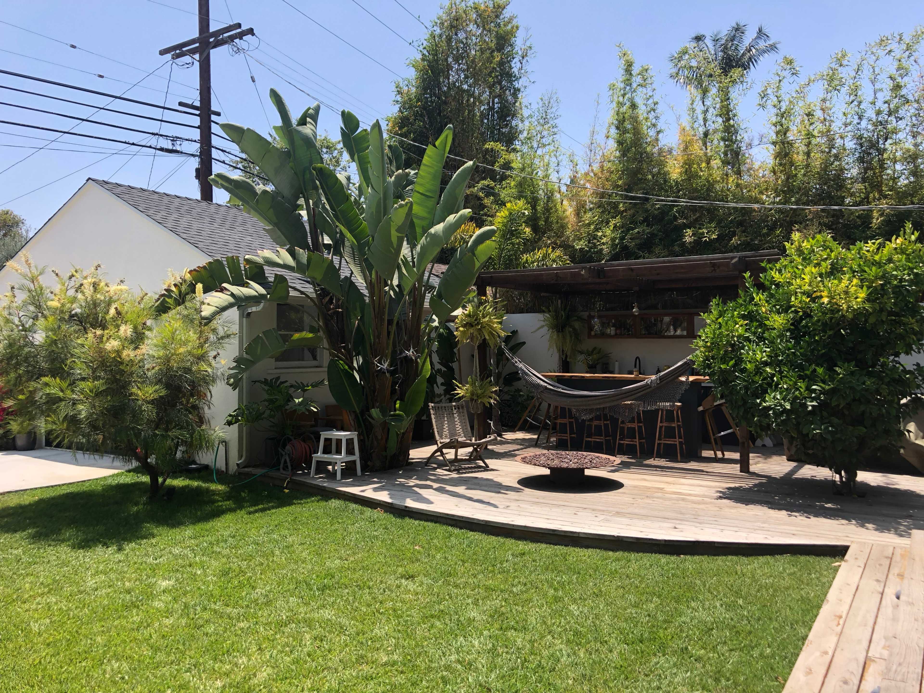 A dwelling with a wooden deck surrounded by lush greenery and large plants, featuring a hammock and outdoor dining area.