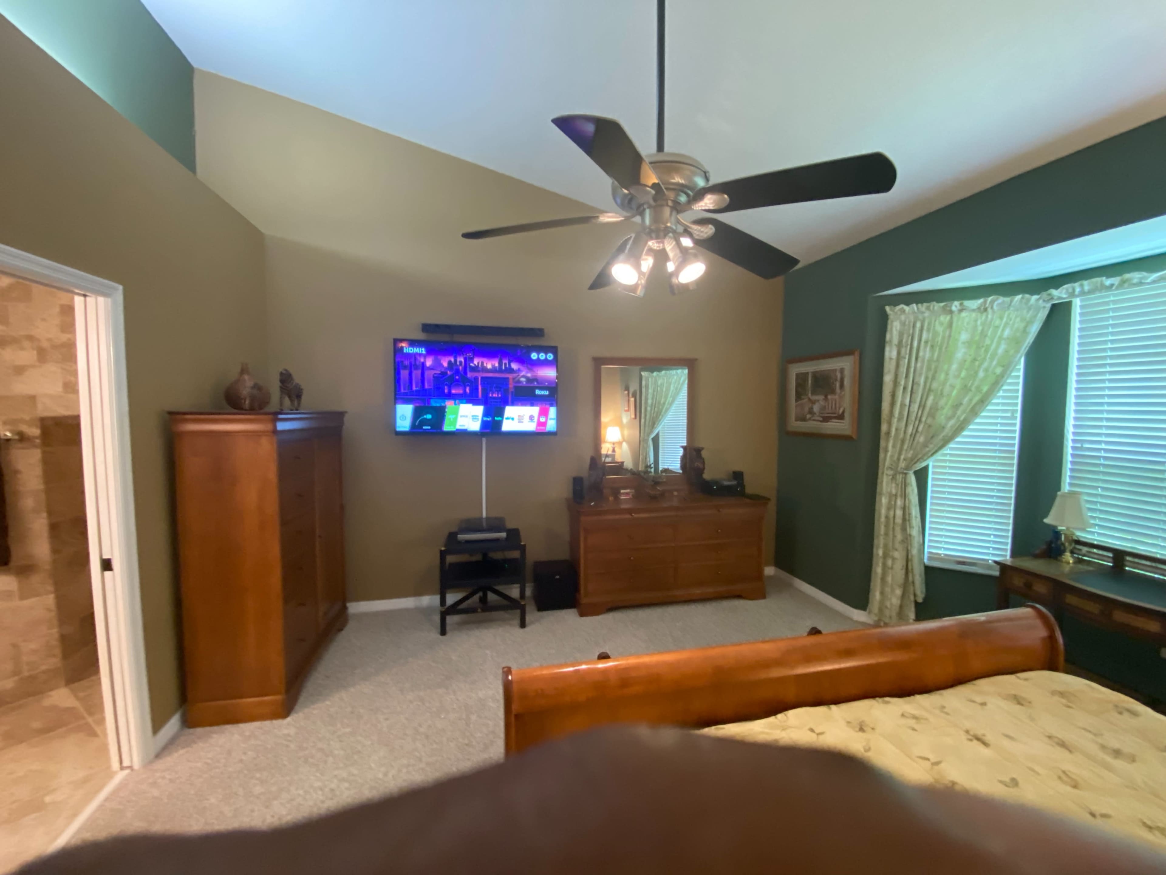 A bedroom features a wooden bed, a dresser, a television mounted on the wall, and windows with curtains, alongside an en-suite bathroom.