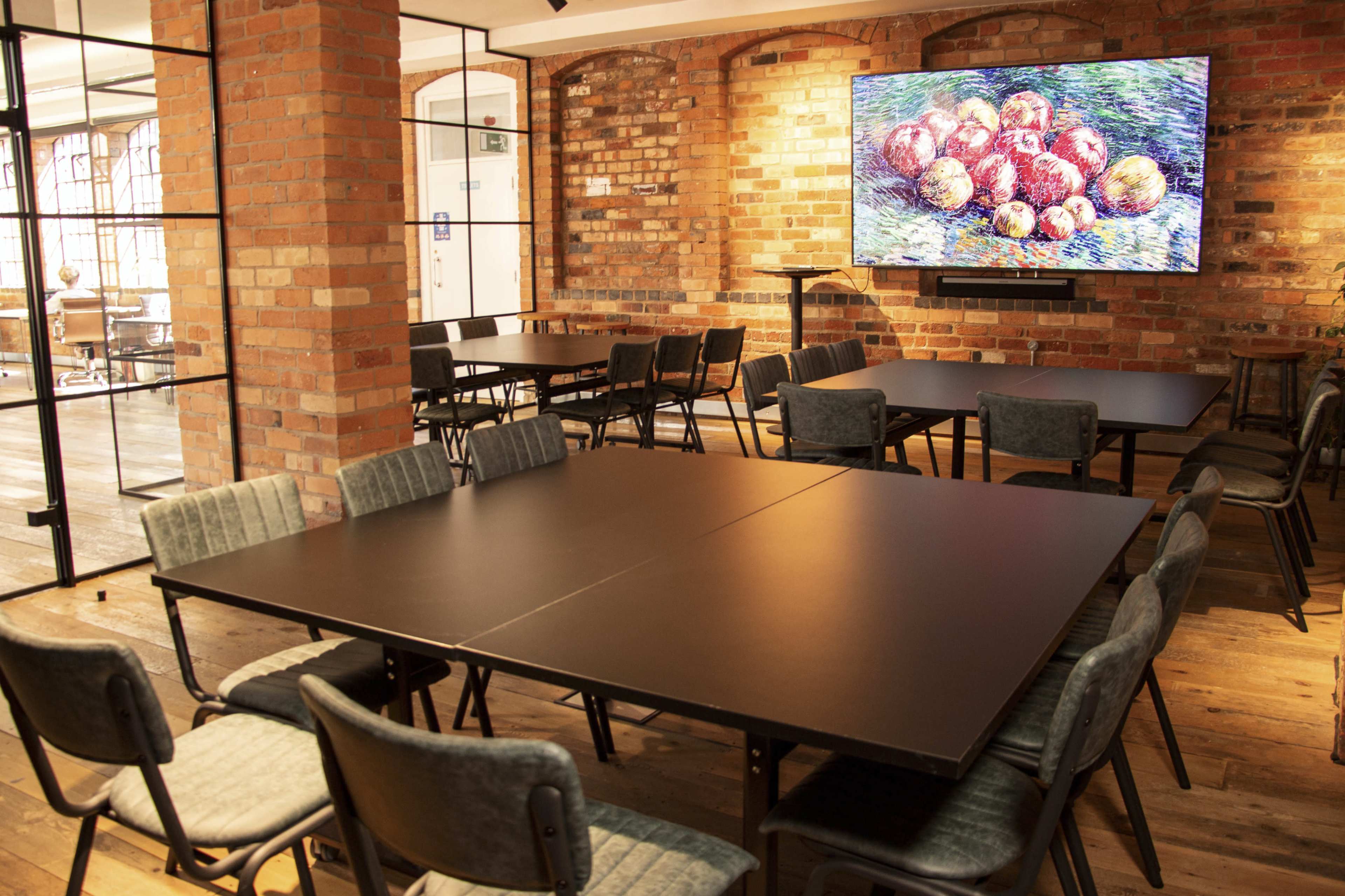 The image shows a modern meeting room with brick walls, several round tables, gray chairs, and a large colorful artwork displayed on the wall.