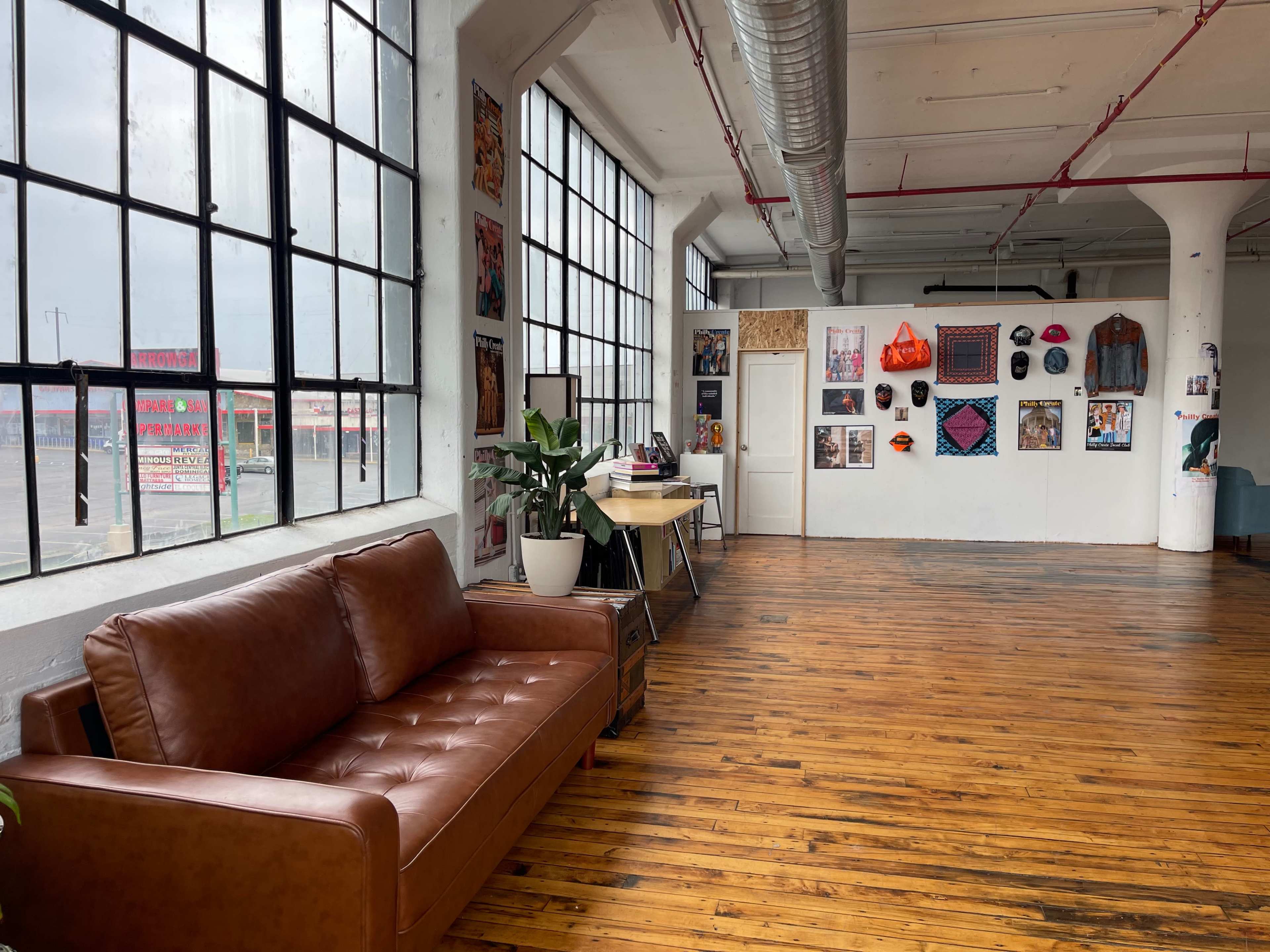 The image shows a spacious room with large windows, featuring a brown leather couch, a desk, and a wall adorned with colorful artwork.