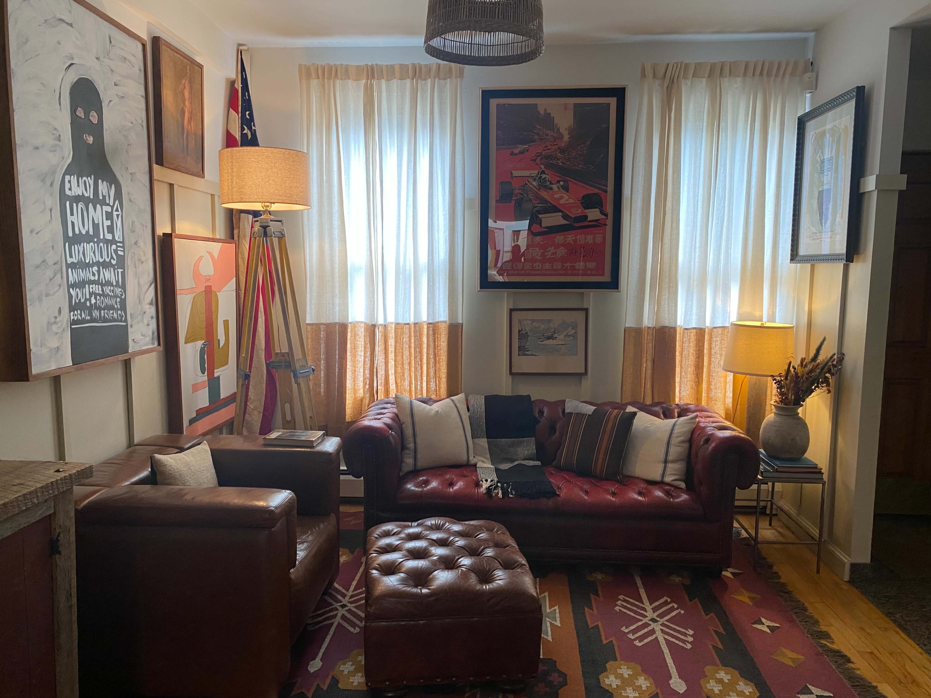 The image shows a cozy living room with a red leather chesterfield sofa, an armchair, an ottoman, and various pieces of art on the walls, illuminated by natural light through window curtains.
