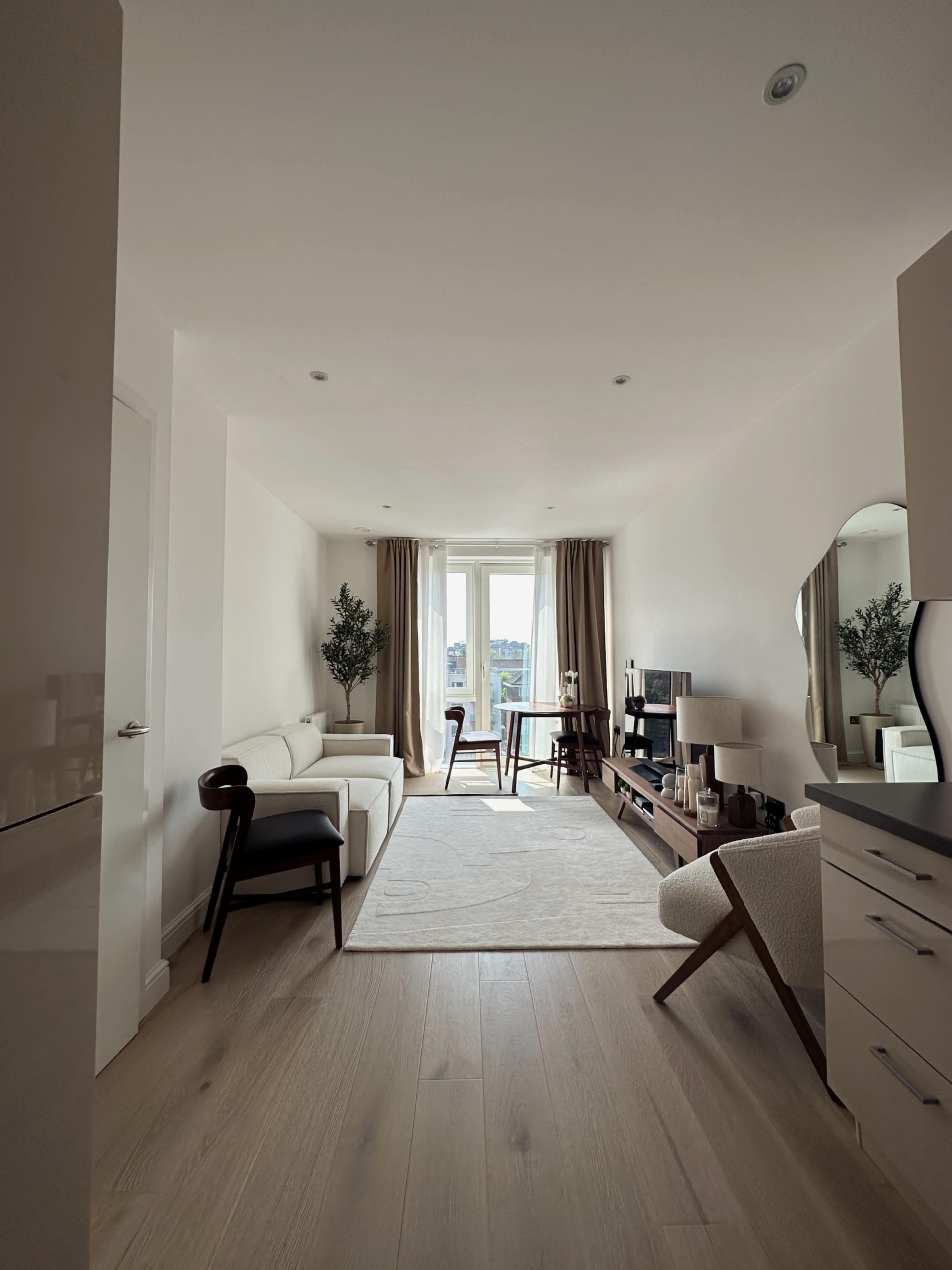 A bright living room features a white sofa, a coffee table, and large windows with beige curtains, alongside decorative plants and modern furniture.