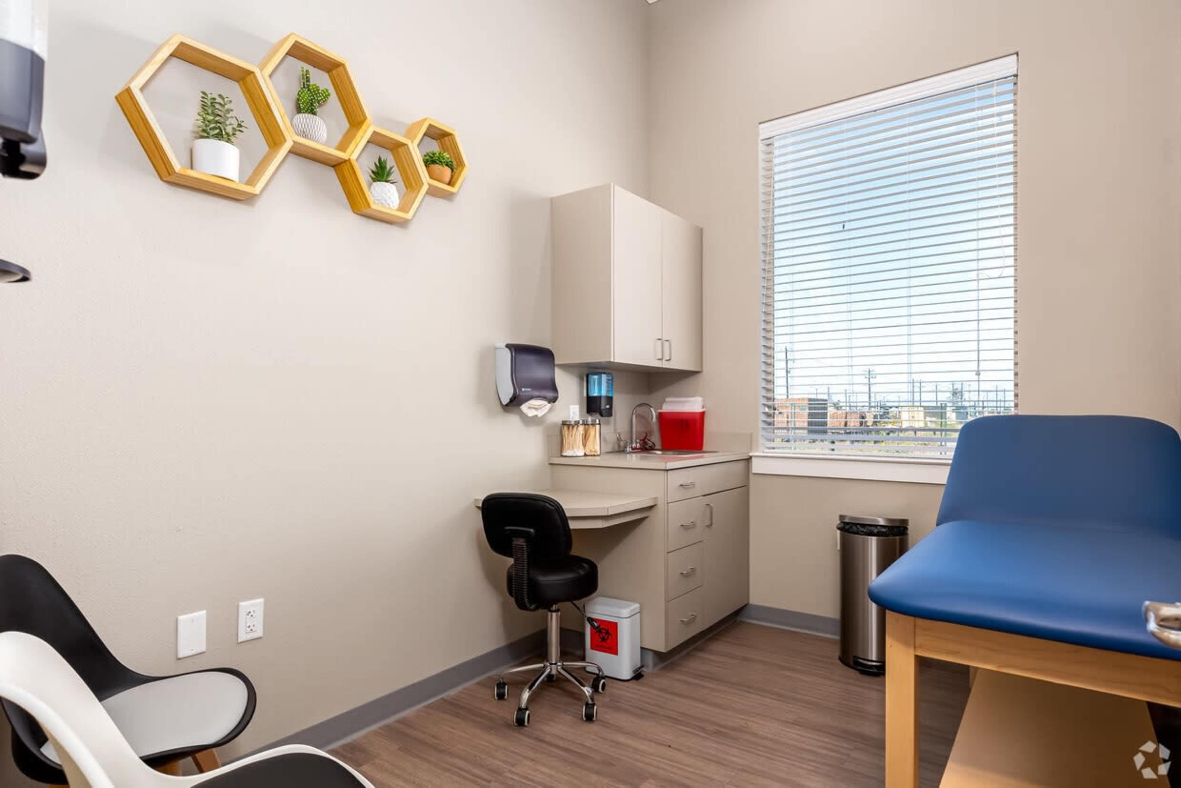 A small examination room featuring a desk with a water dispenser, a chair, a medical examination table, and decorative hexagonal shelves with plants.