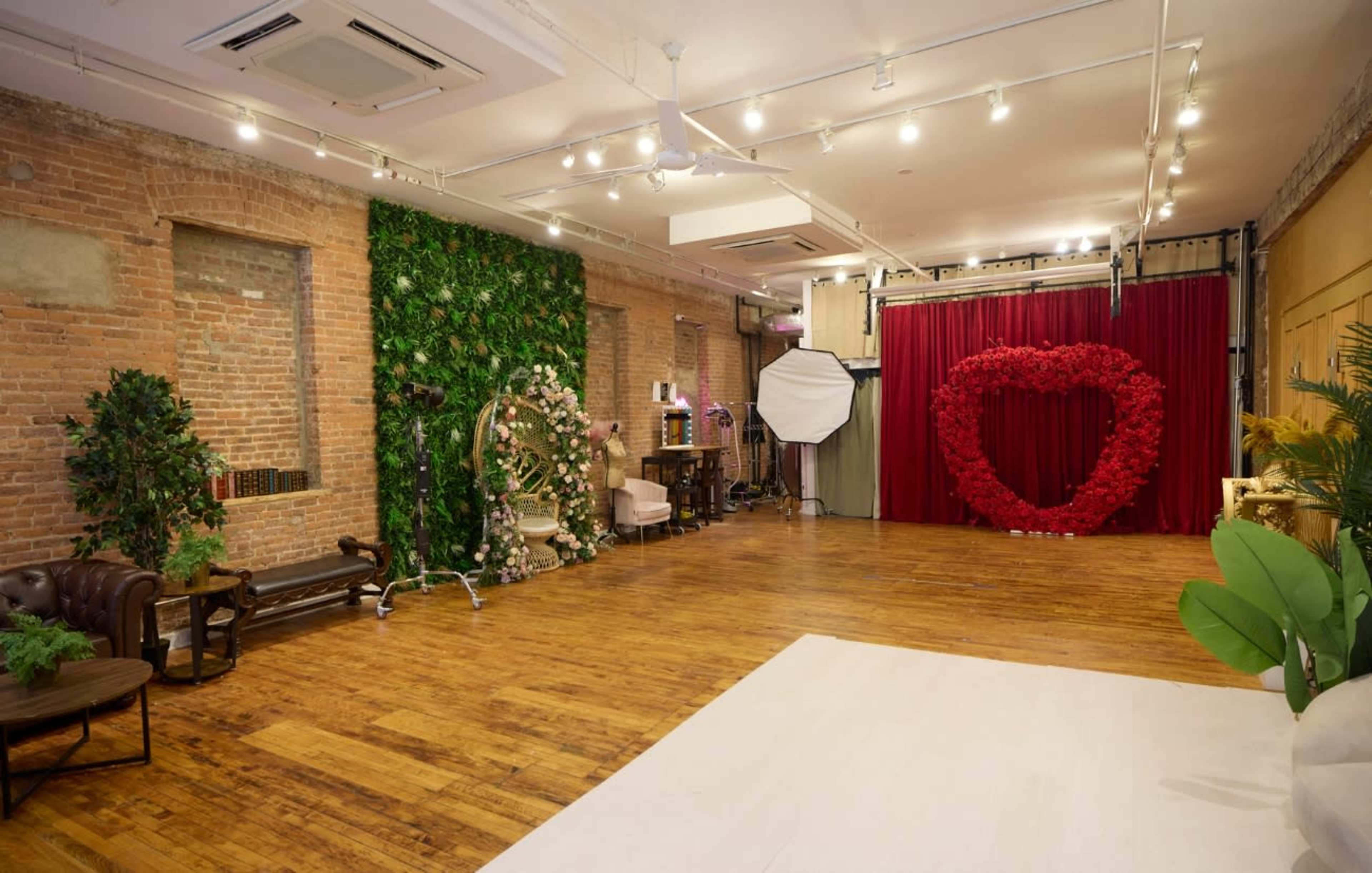 The image features a spacious studio with brick walls, a wooden floor, and decorated areas including a green plant wall, a floral heart backdrop, and various furniture pieces.