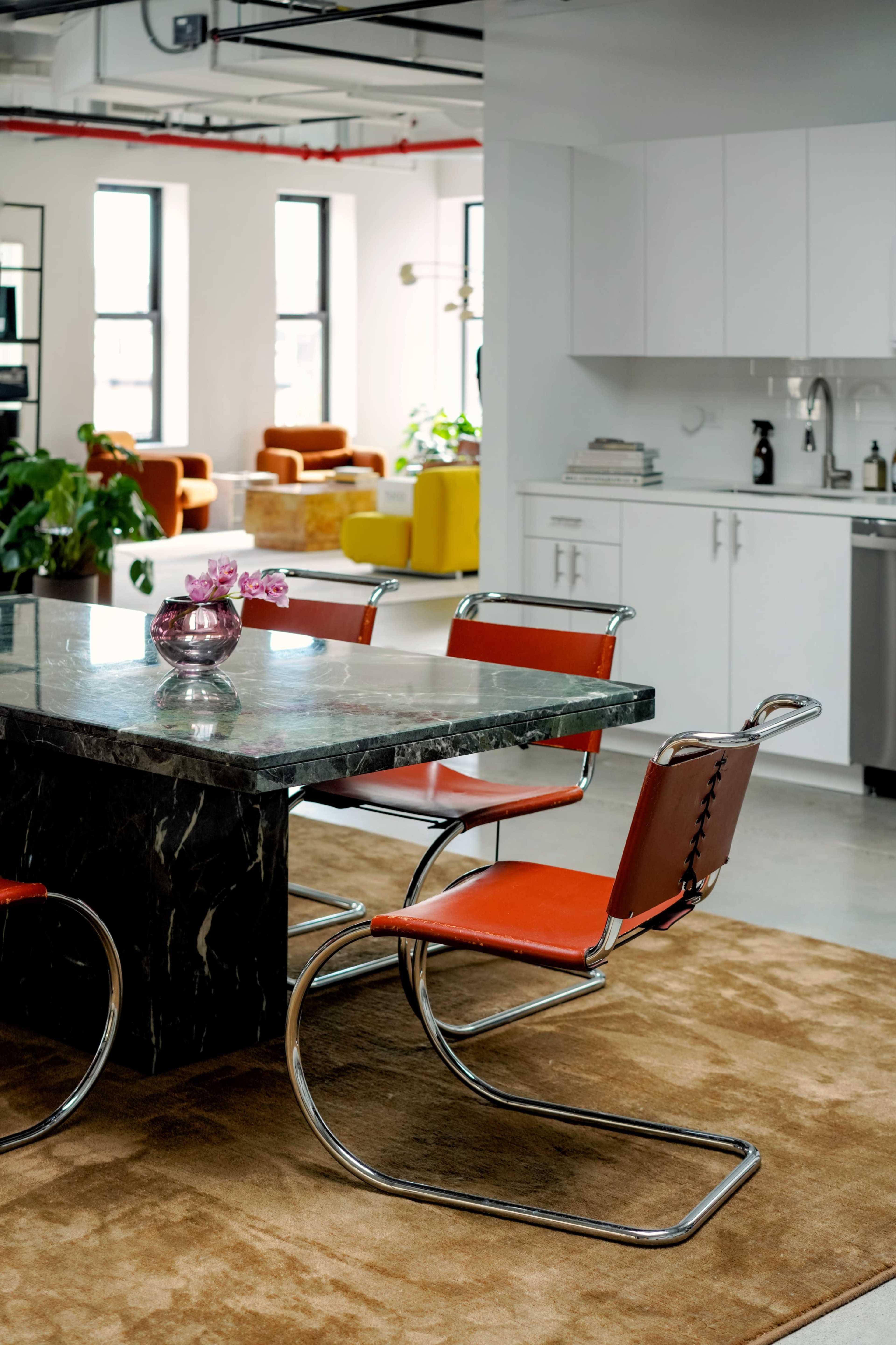 A modern dining area features a marble table with orange chairs and a vase of flowers, surrounded by a light-filled kitchen and additional seating.