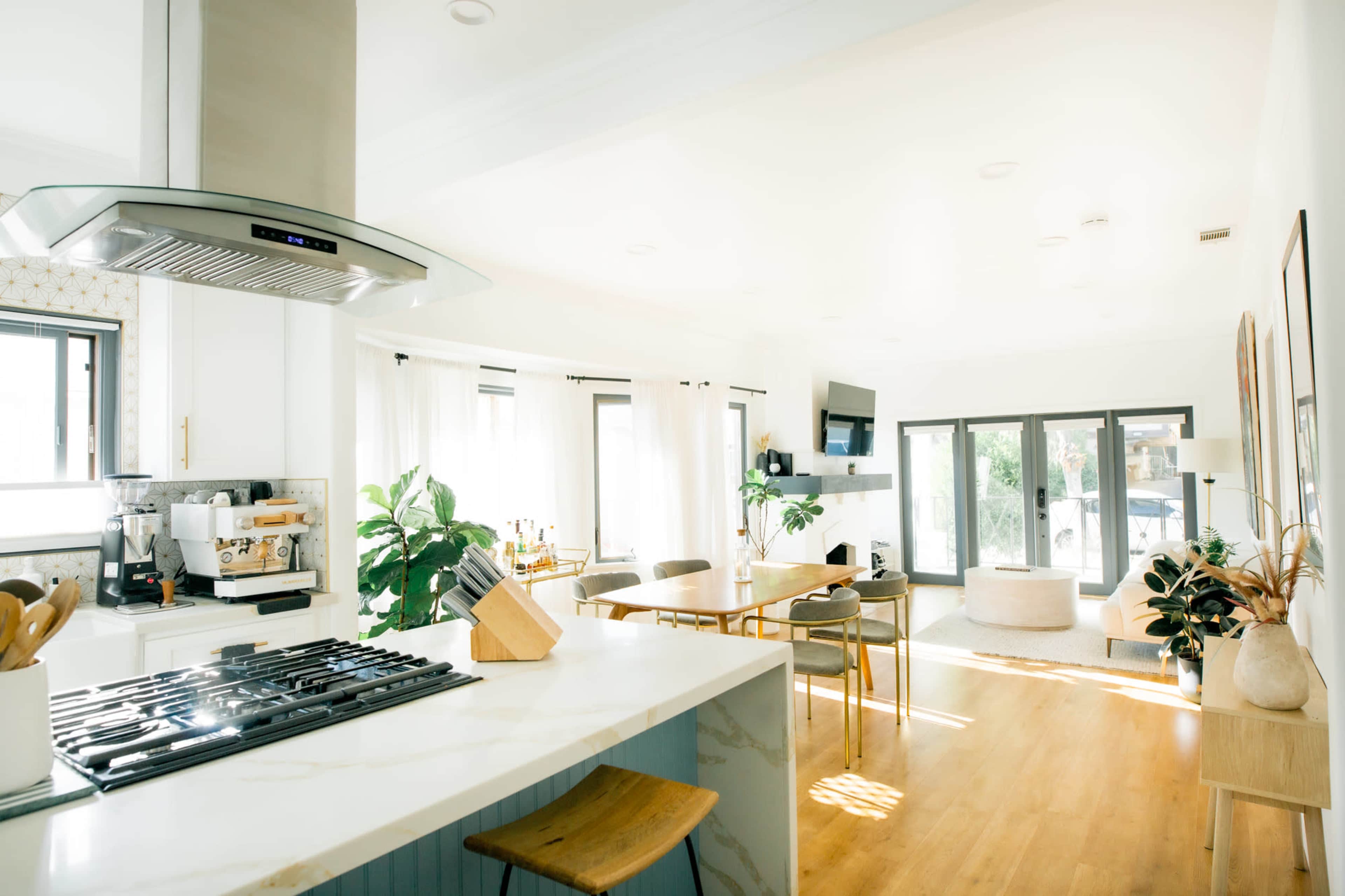 A bright and open kitchen features a marble countertop, modern appliances, and a dining area adjacent to a spacious living room with large windows.