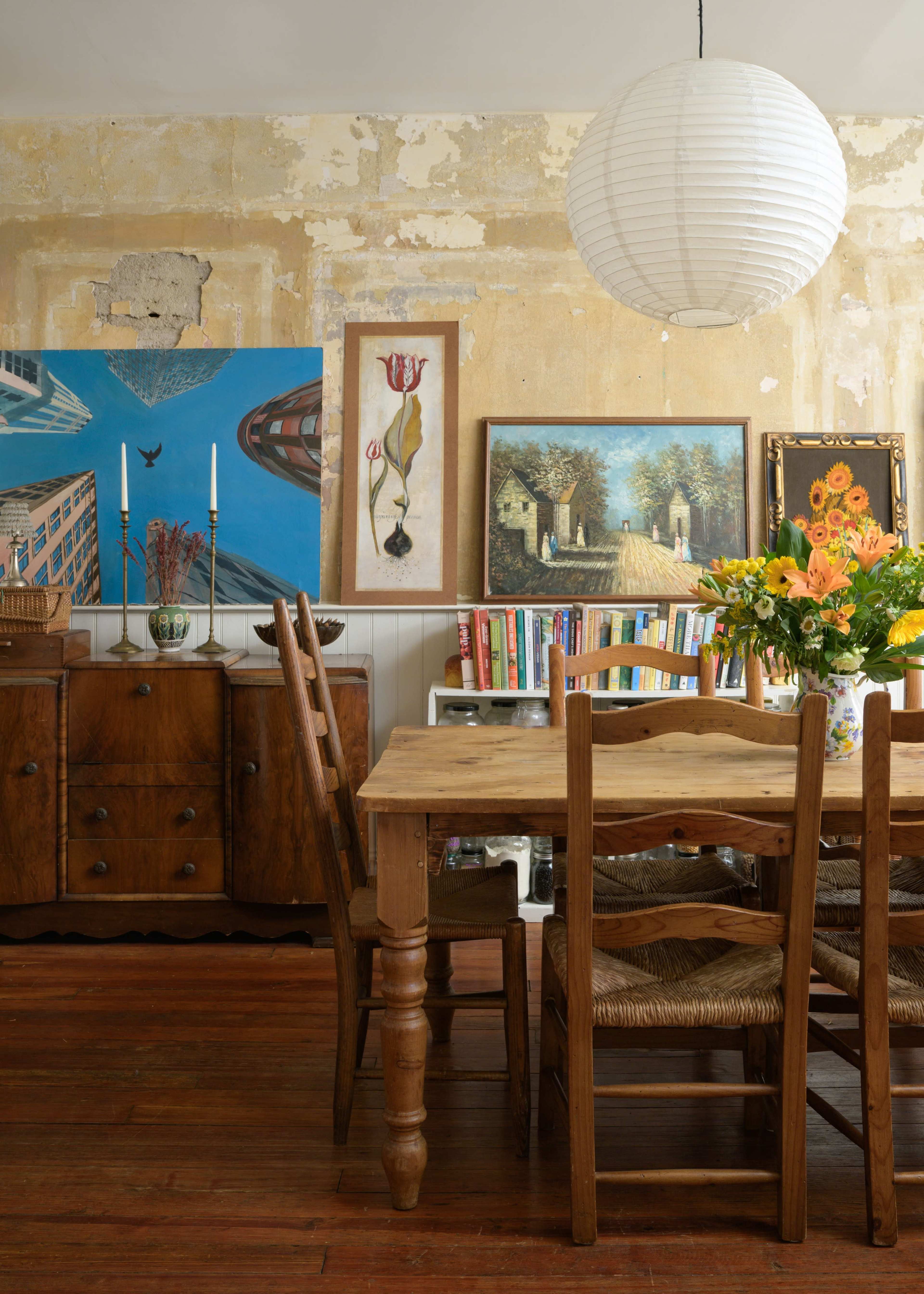 A wooden dining table surrounded by chairs is set against a wall featuring various framed artworks and a round paper lantern overhead.