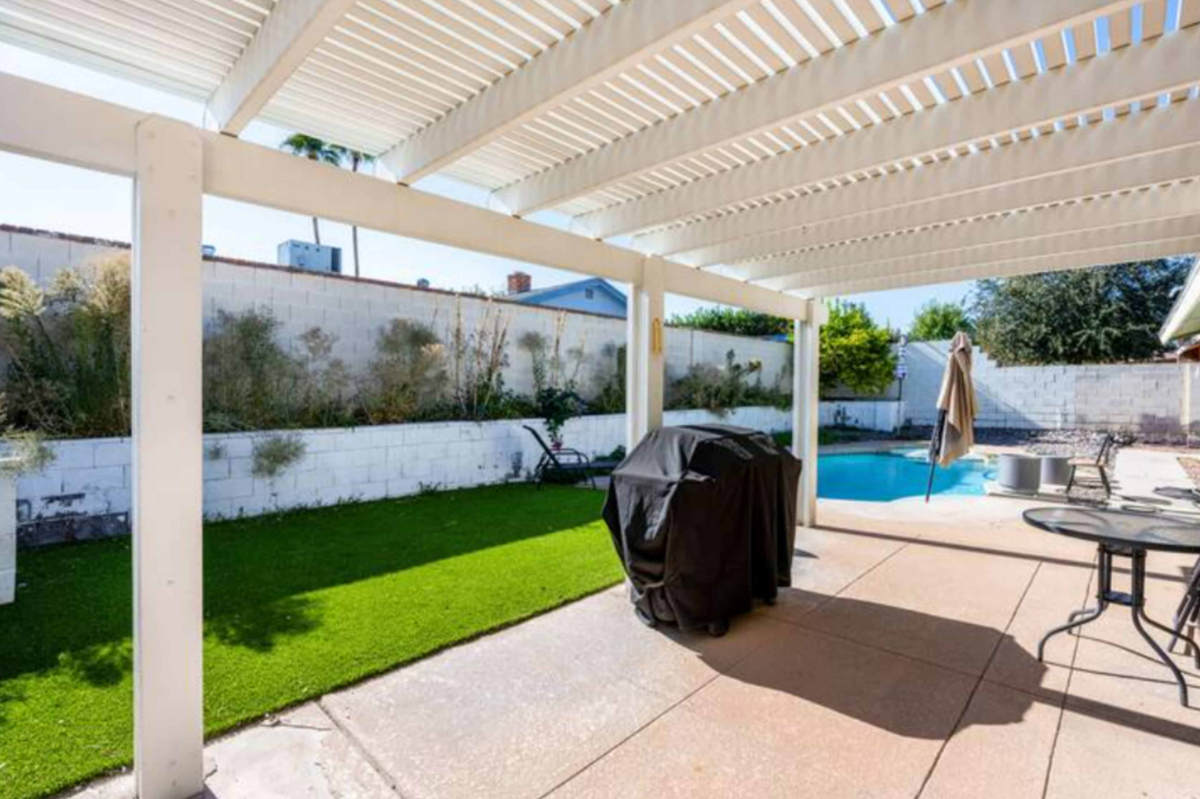 A covered patio features a pool and a grill, surrounded by a grassy area and a white wall.