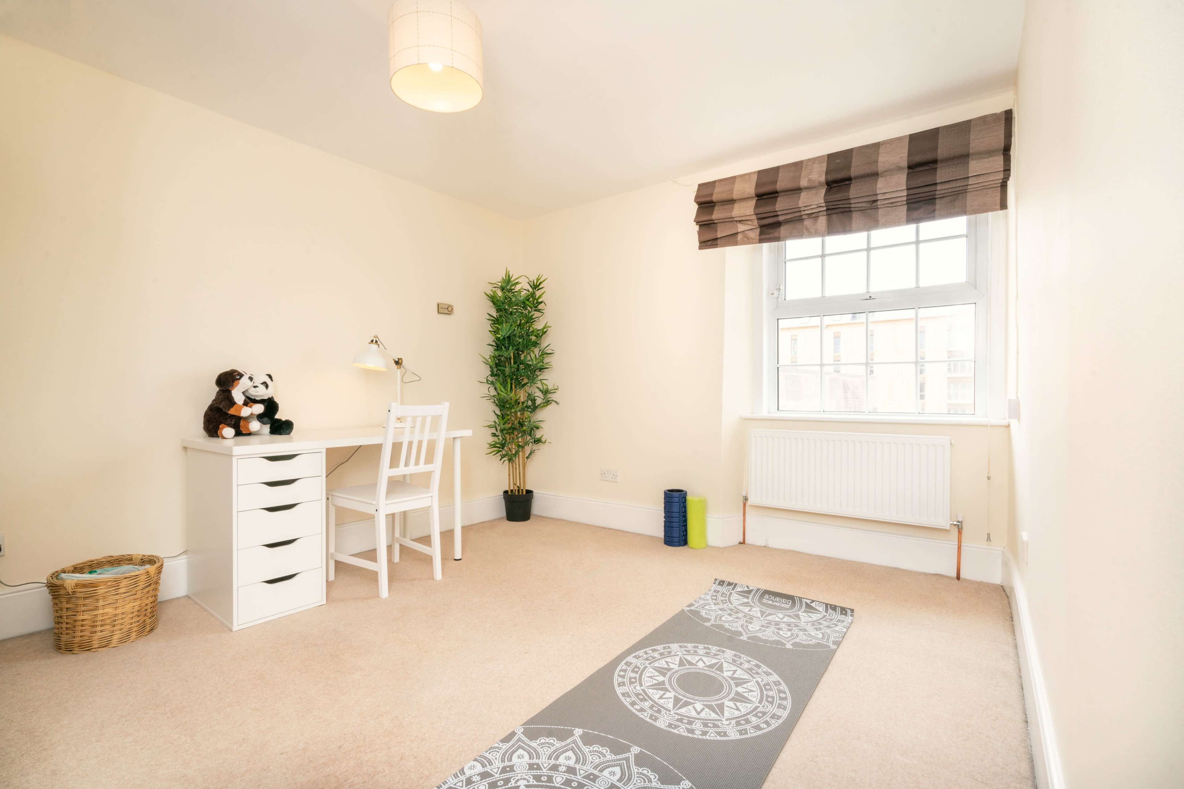A minimalistic room with a desk and chair near a window, a plant in the corner, and a yoga mat on the floor.