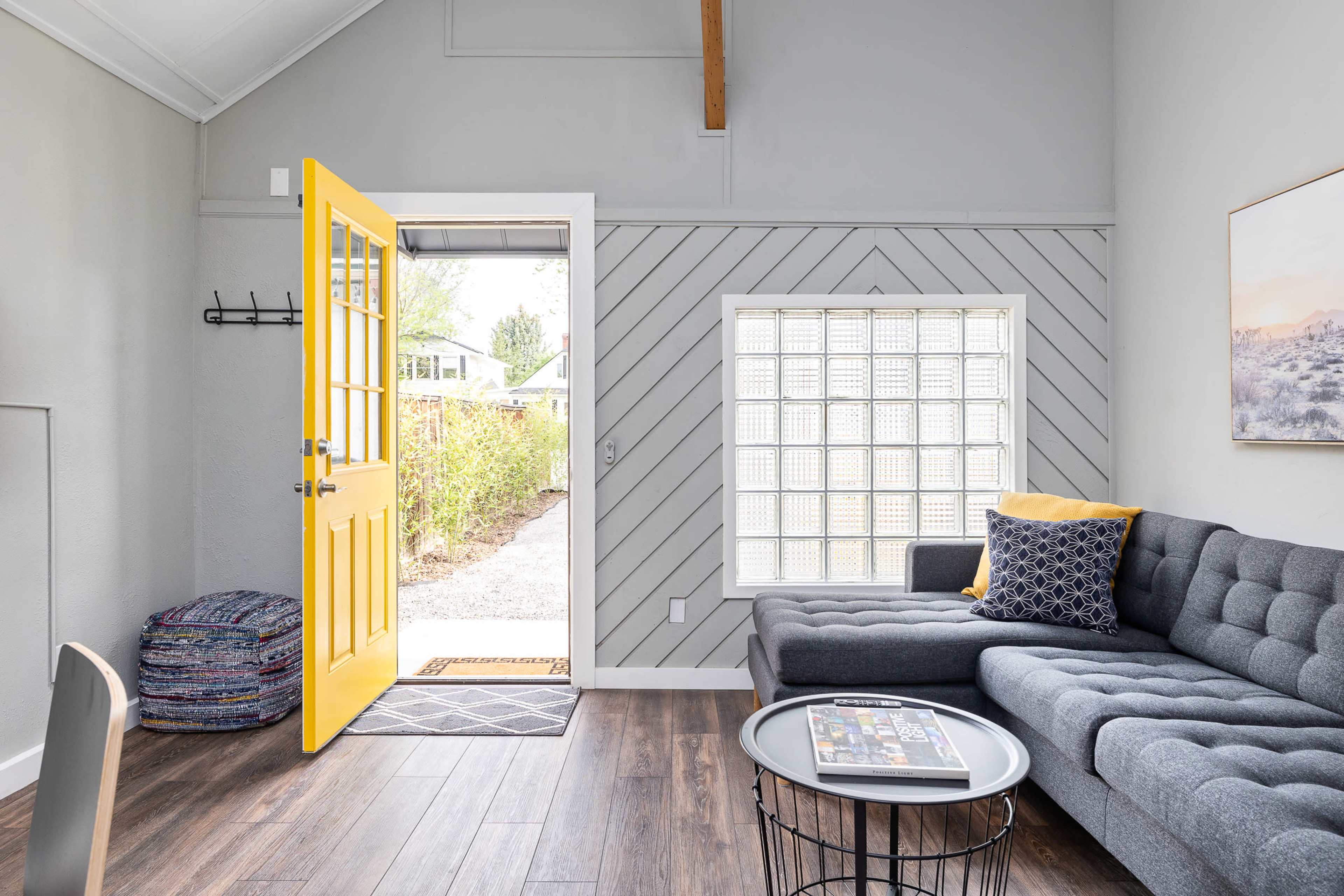 A bright yellow front door opens into a modern living room with a gray couch, a glass block window, and natural light streaming in.