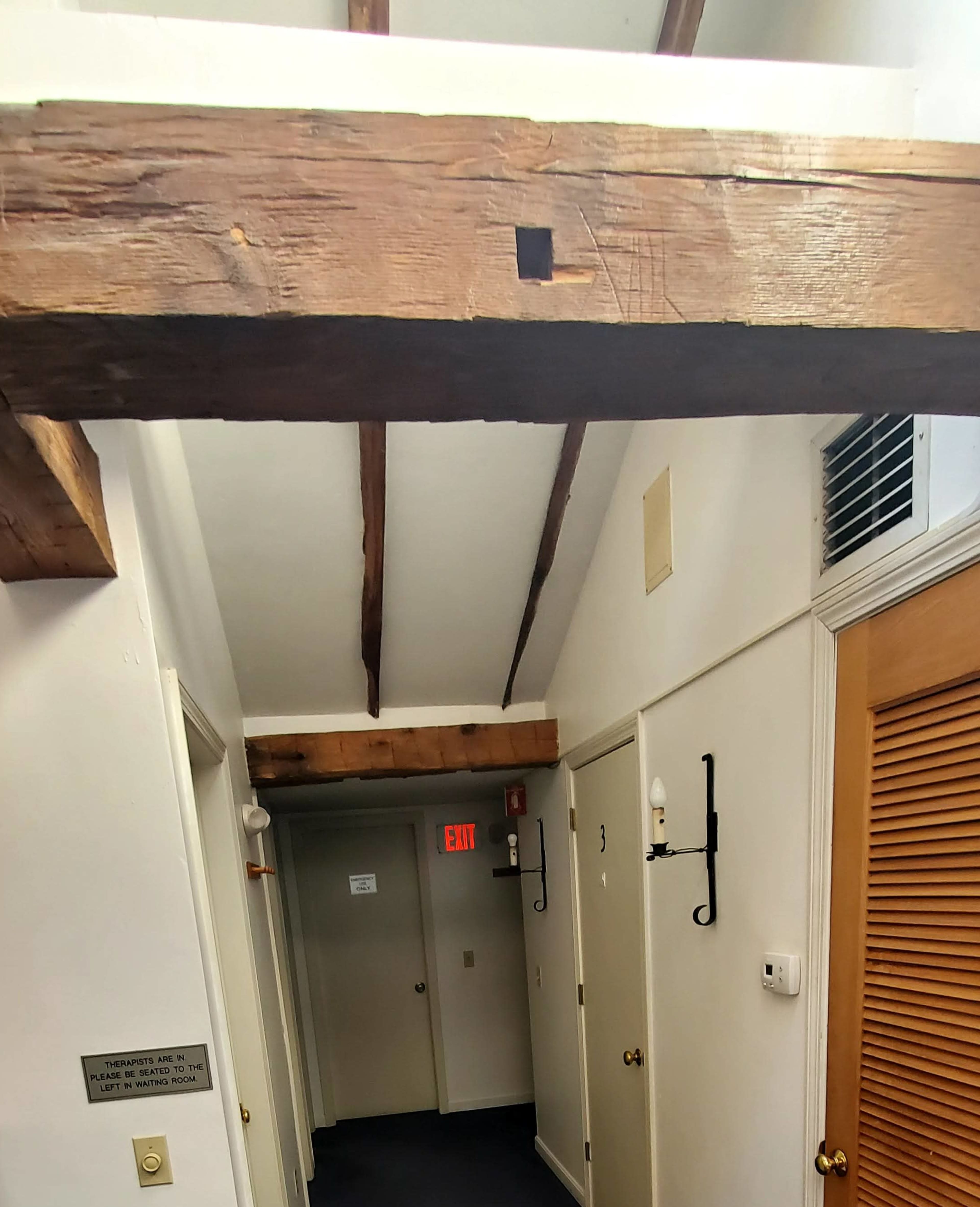 A narrow hallway with wooden beams on the ceiling, light-colored walls, and several doors along the sides.