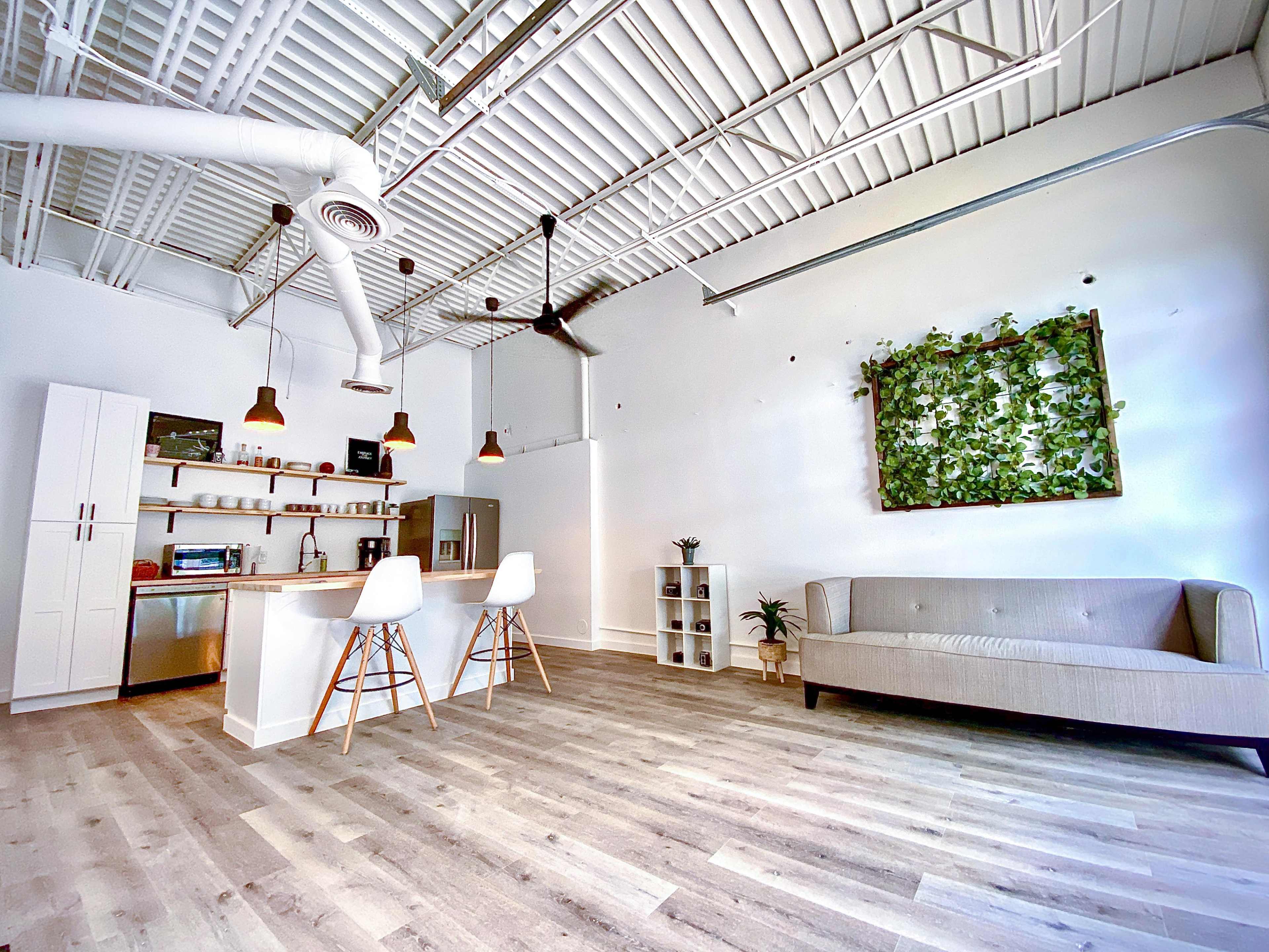 A spacious, modern interior with a kitchenette, a couch, and a green wall decoration featuring plants.
