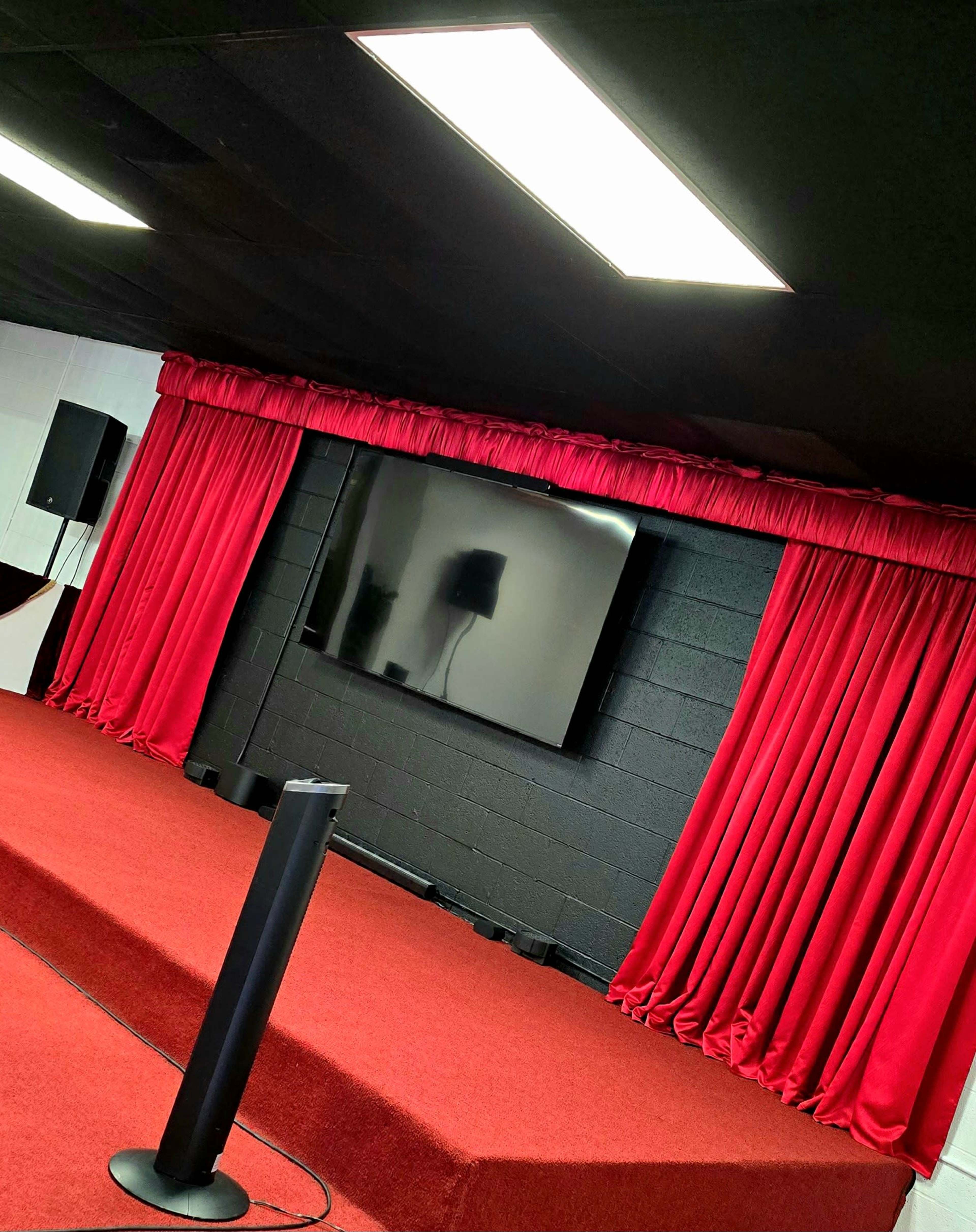 The image shows a stage area with red curtains, a large flat-screen TV mounted on a black wall, and a red carpeted floor.