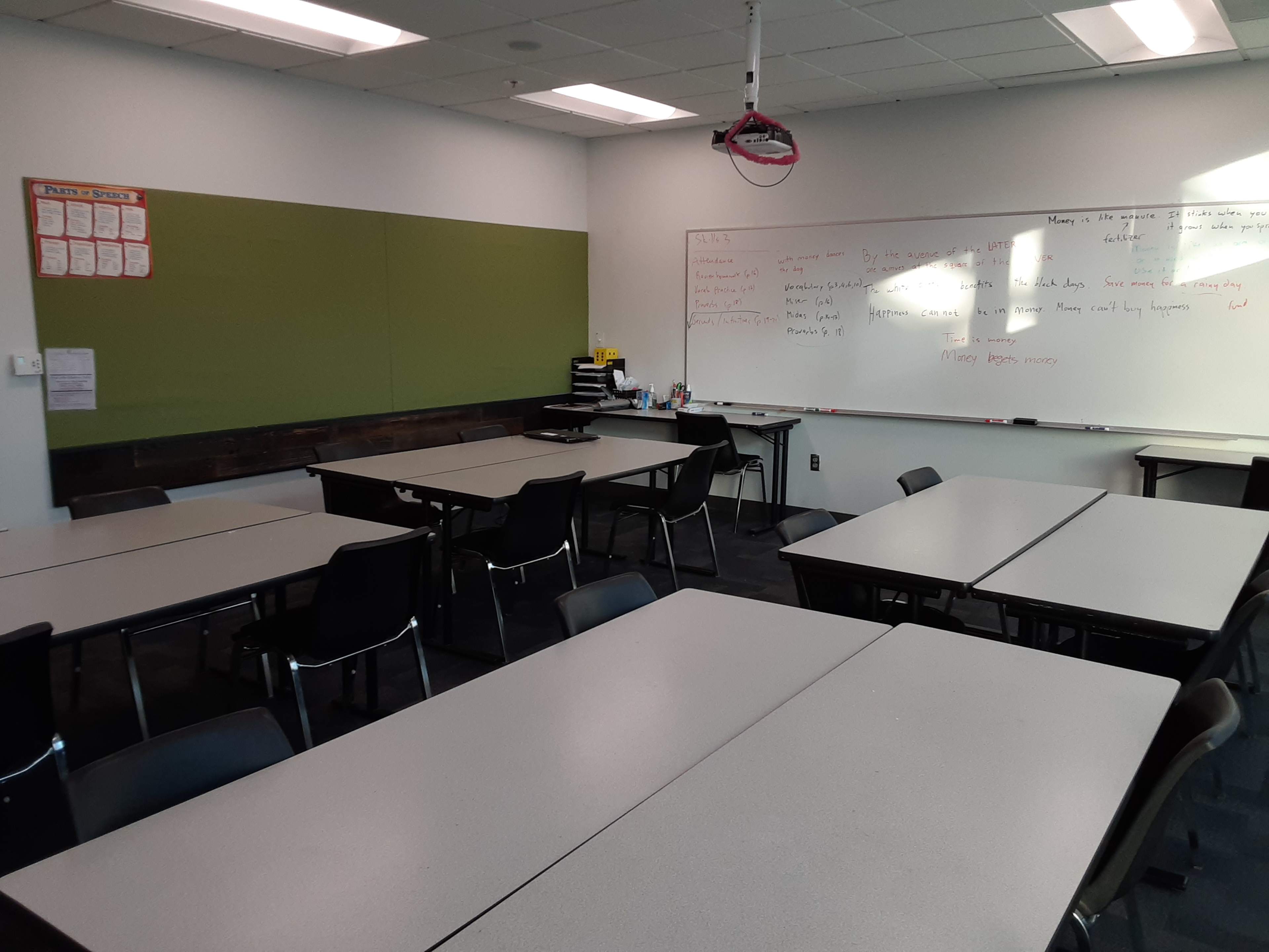 A classroom is set up with several tables and chairs arranged around a whiteboard and a green wall.