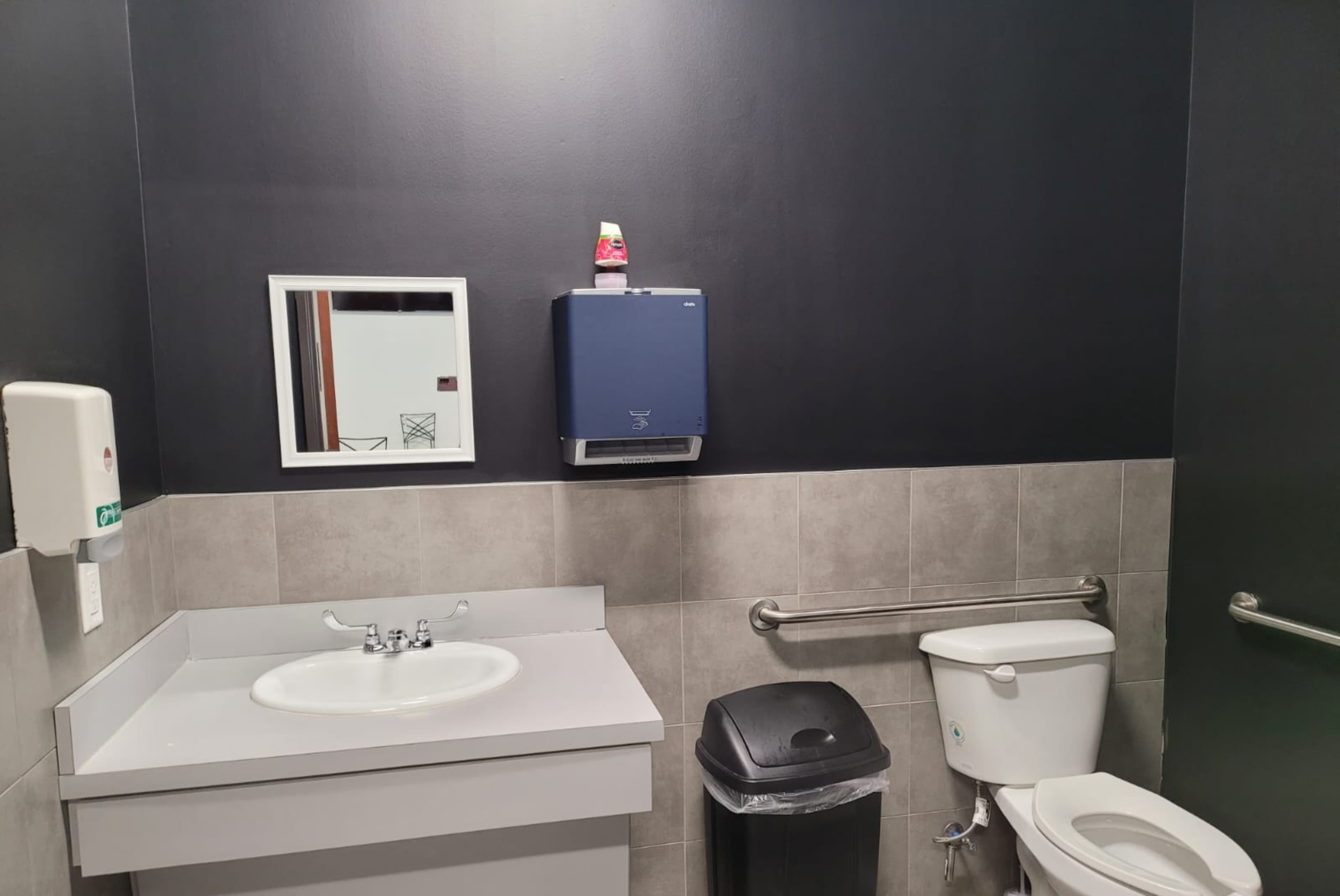 A bathroom with a sink, toilet, mirror, and a hand dryer against a dark wall.