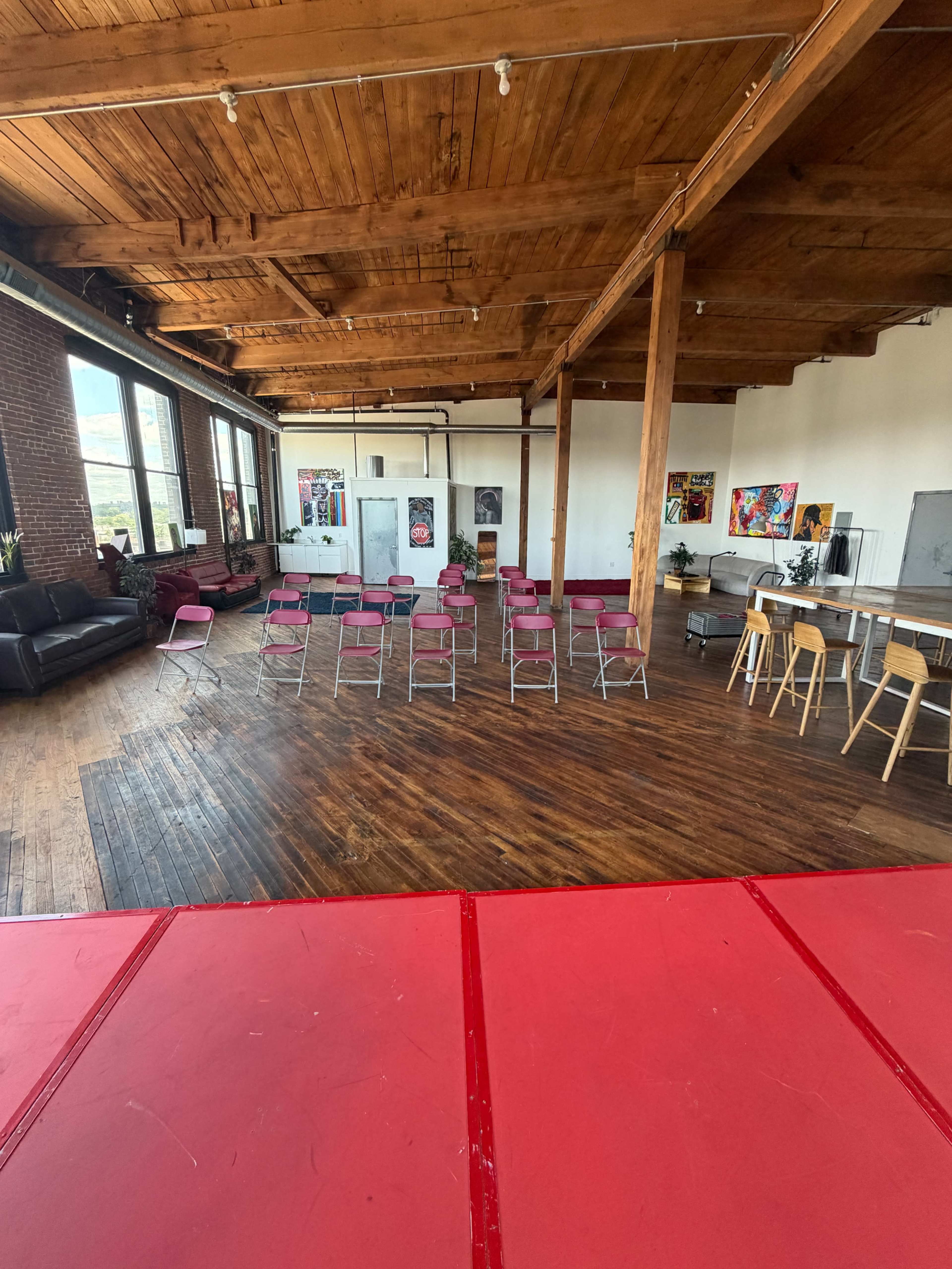 The image shows a spacious studio or event space with wooden beams, rows of pink folding chairs set up for an audience, and various colorful artworks on the walls.