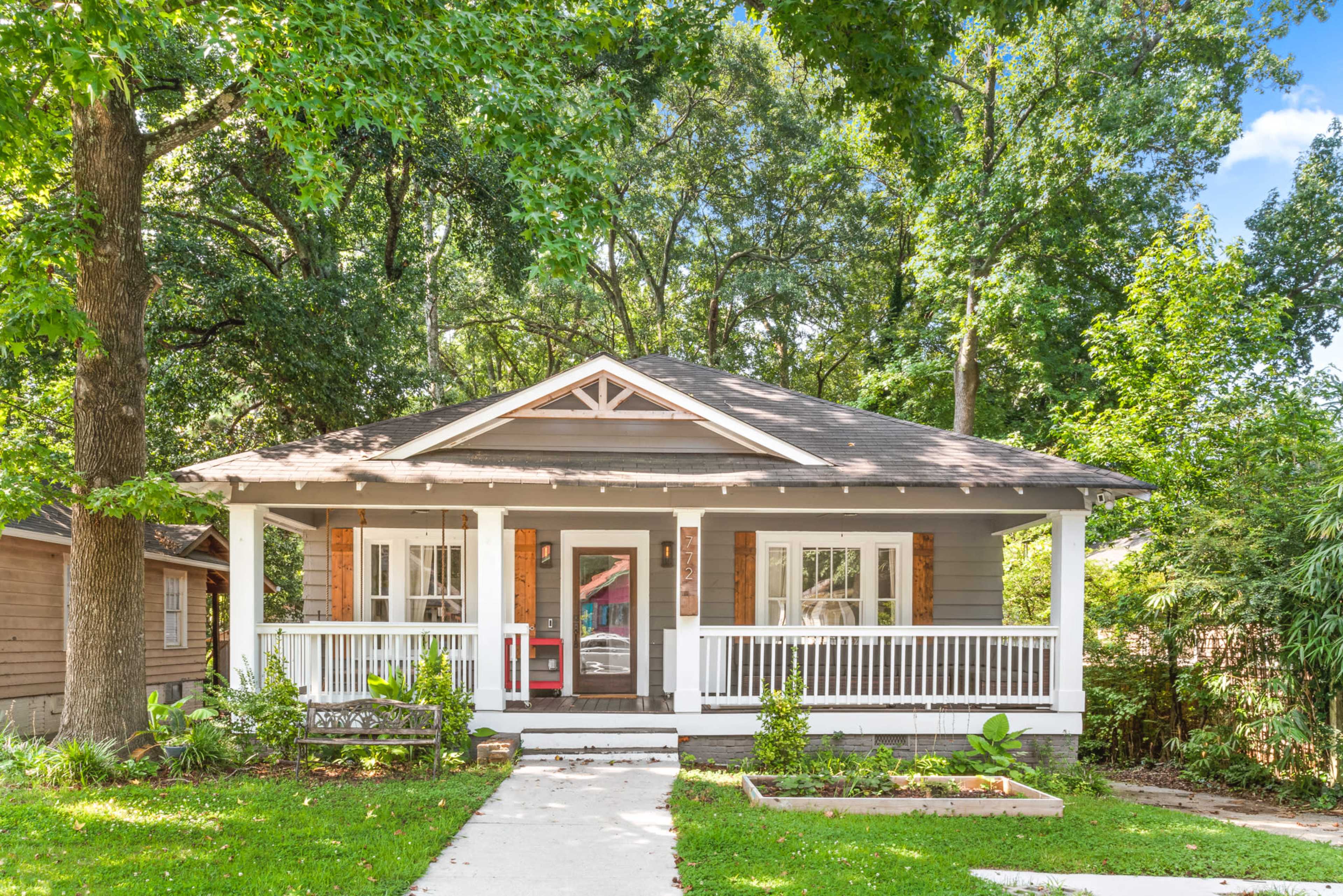 A single-story gray house with a front porch, surrounded by lush green trees and plants.