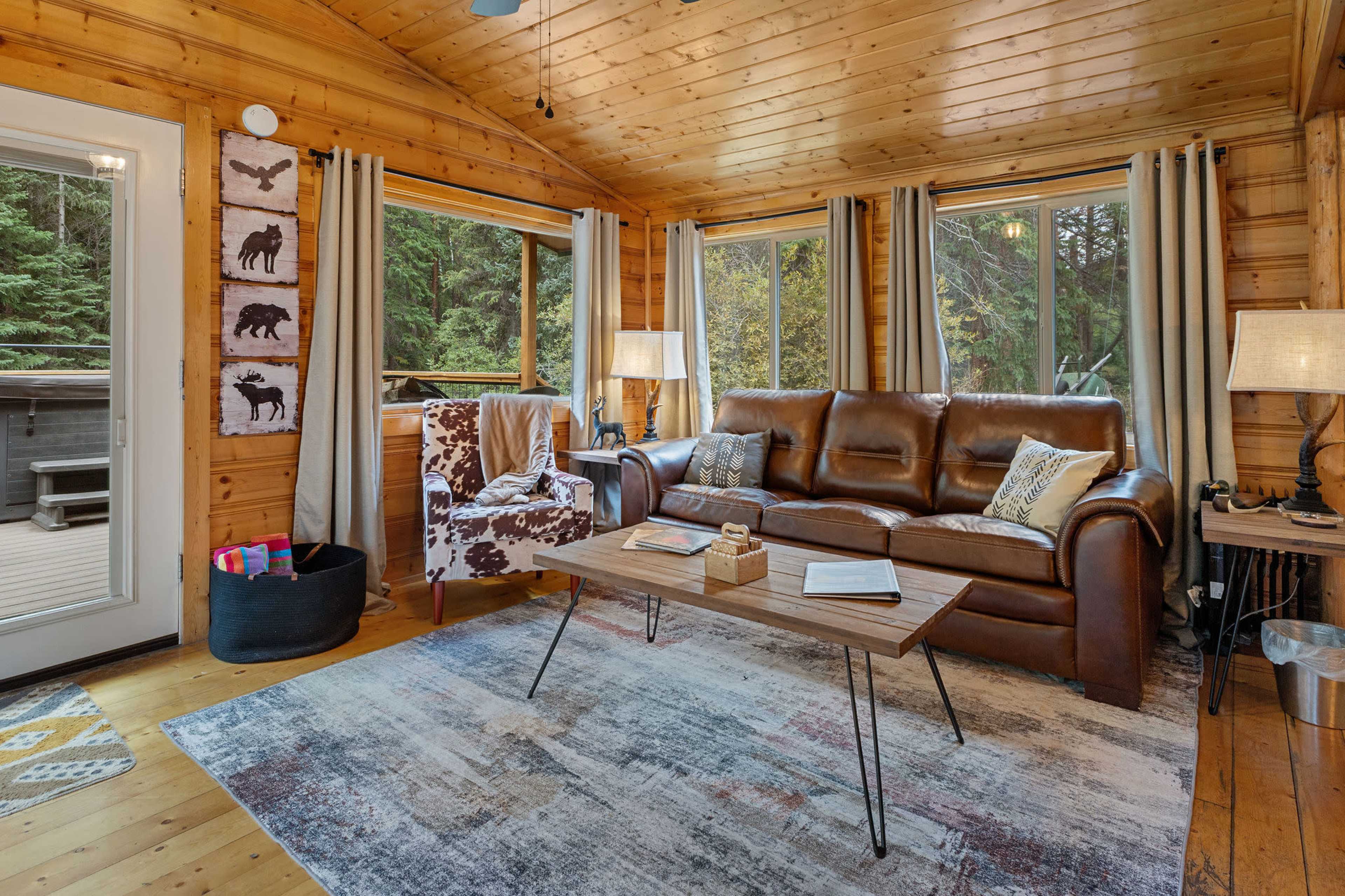 The room features a wooden interior with large windows, a brown leather sofa, a coffee table, and a patterned rug.