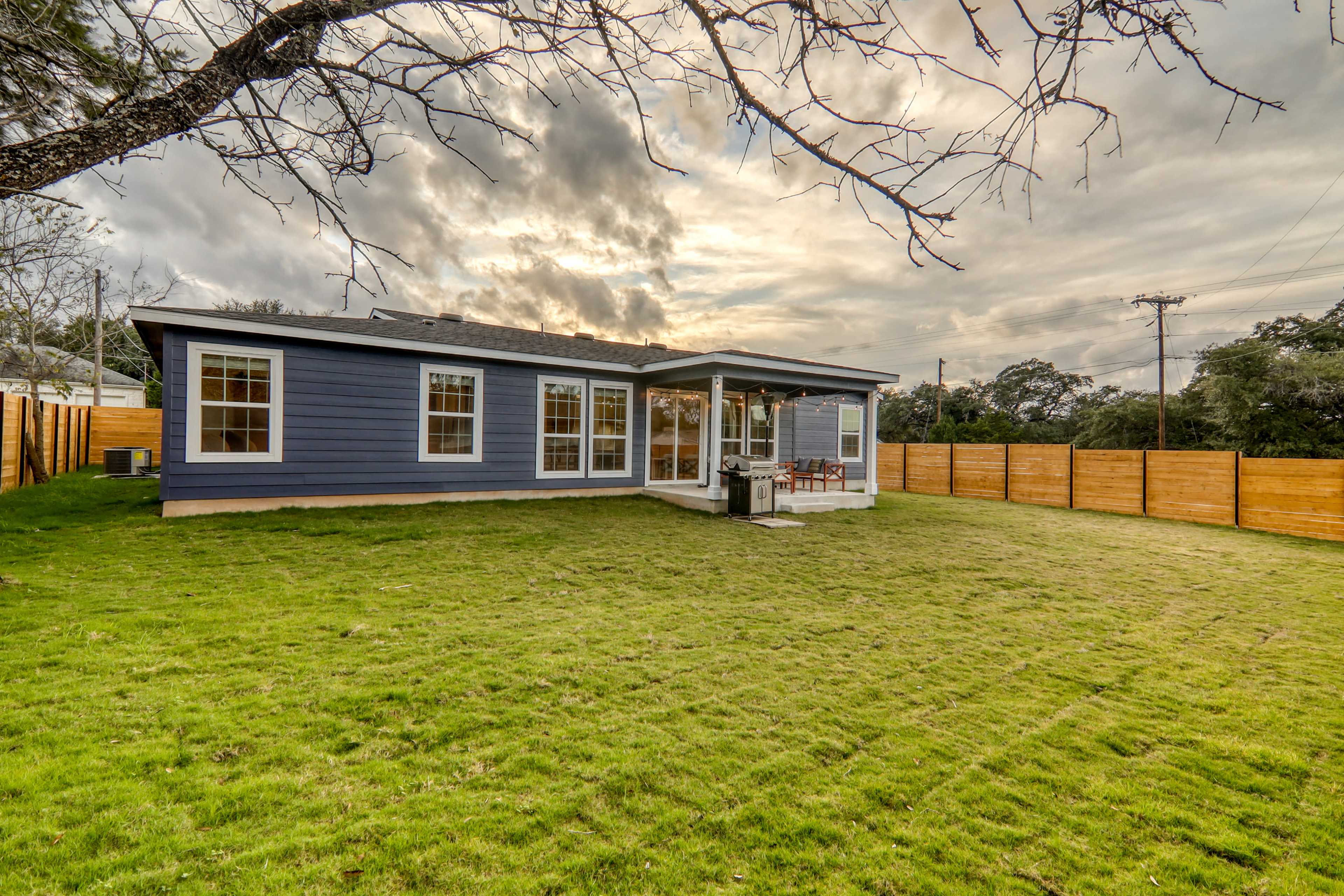 A blue house with large windows and a porch is set in a grassy yard surrounded by wooden fencing under a cloudy sky.