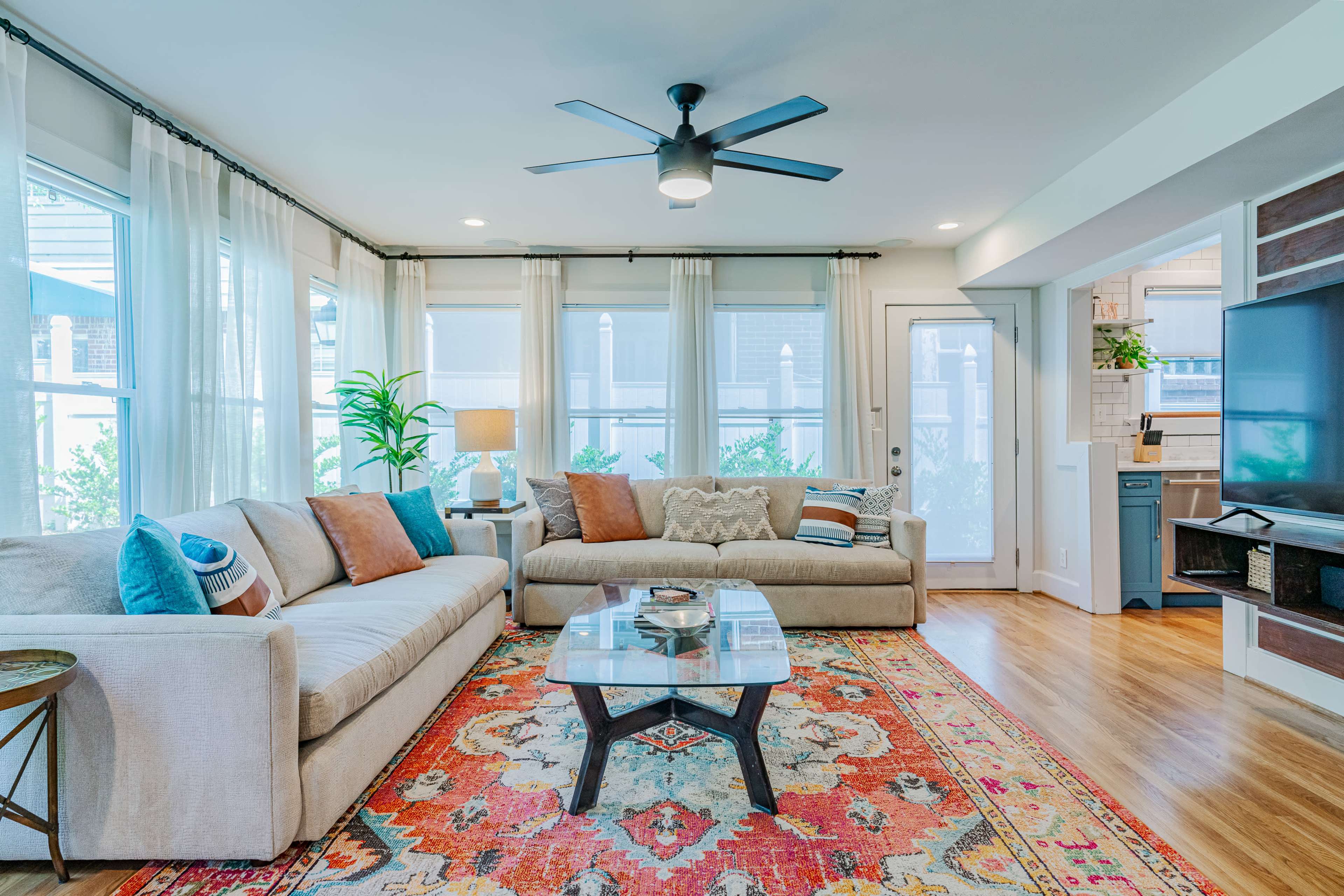 A modern living room featuring a light-colored sofa, a patterned area rug, a ceiling fan, and large windows with sheer curtains.