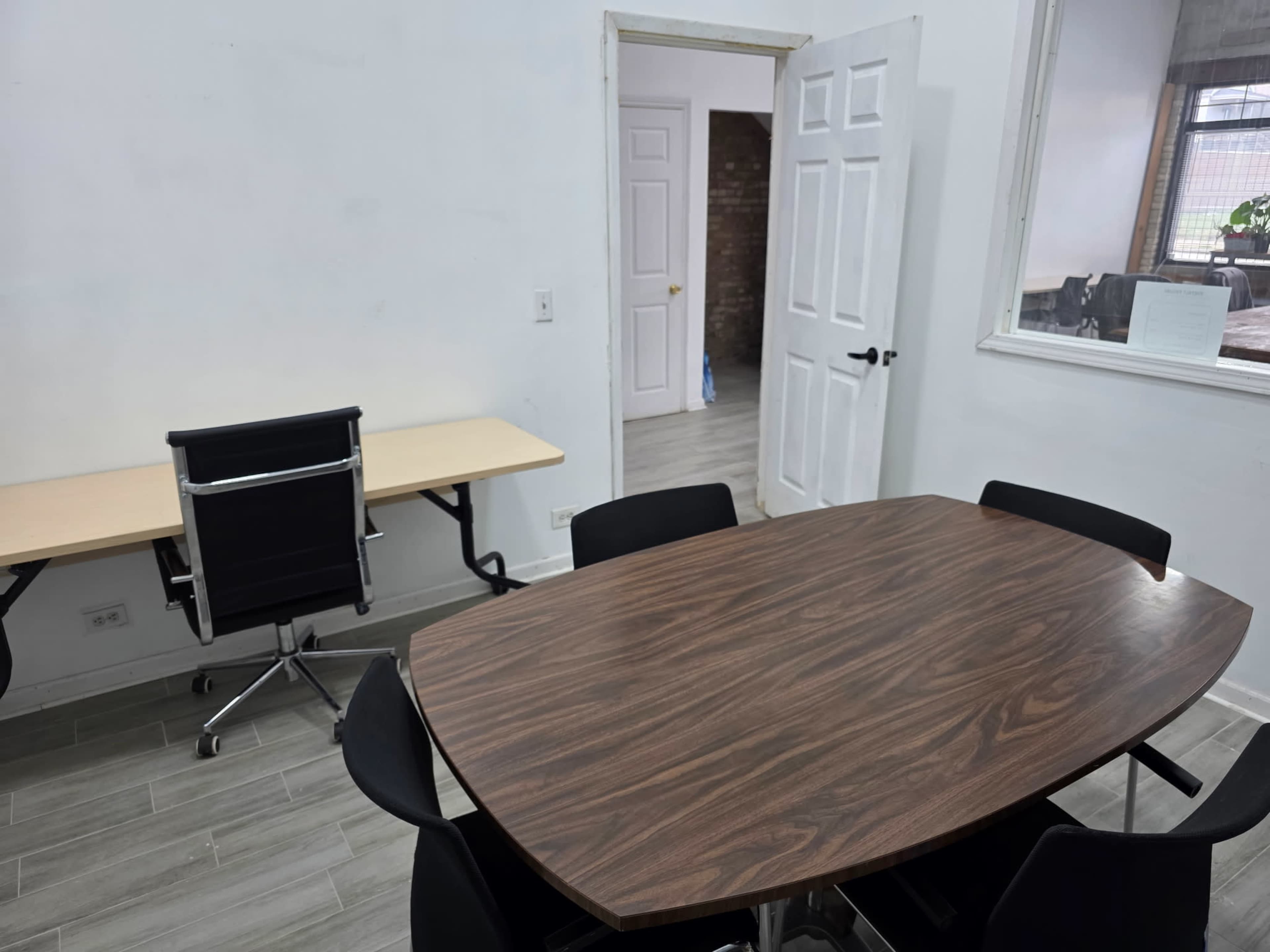 The image shows a minimalist office space with a wooden oval table surrounded by black chairs, a desk with a chair in the background, and an open door leading to another room.
