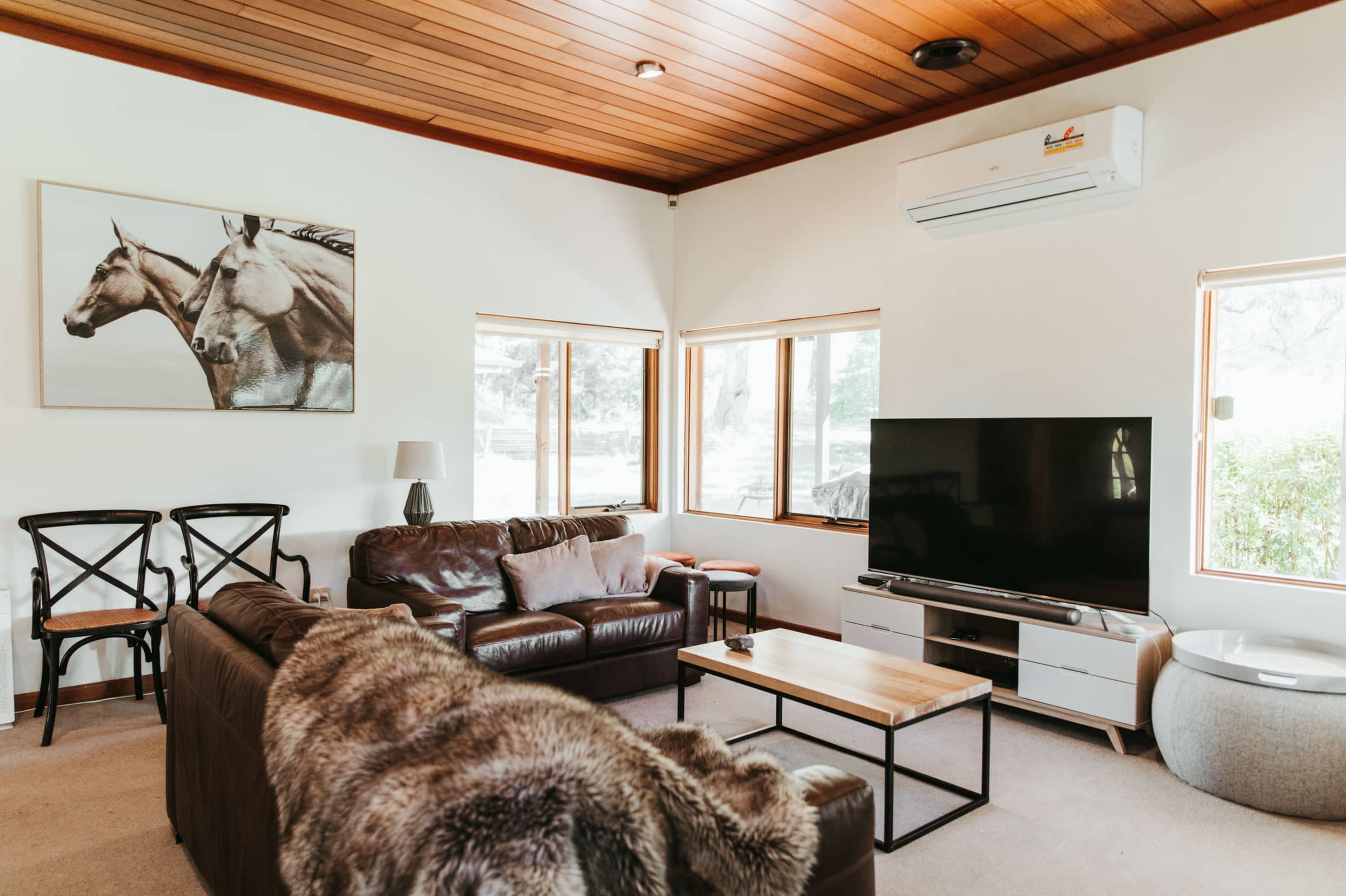 A cozy living room with leather sofas, a wooden coffee table, a large television, and large windows allowing natural light in.