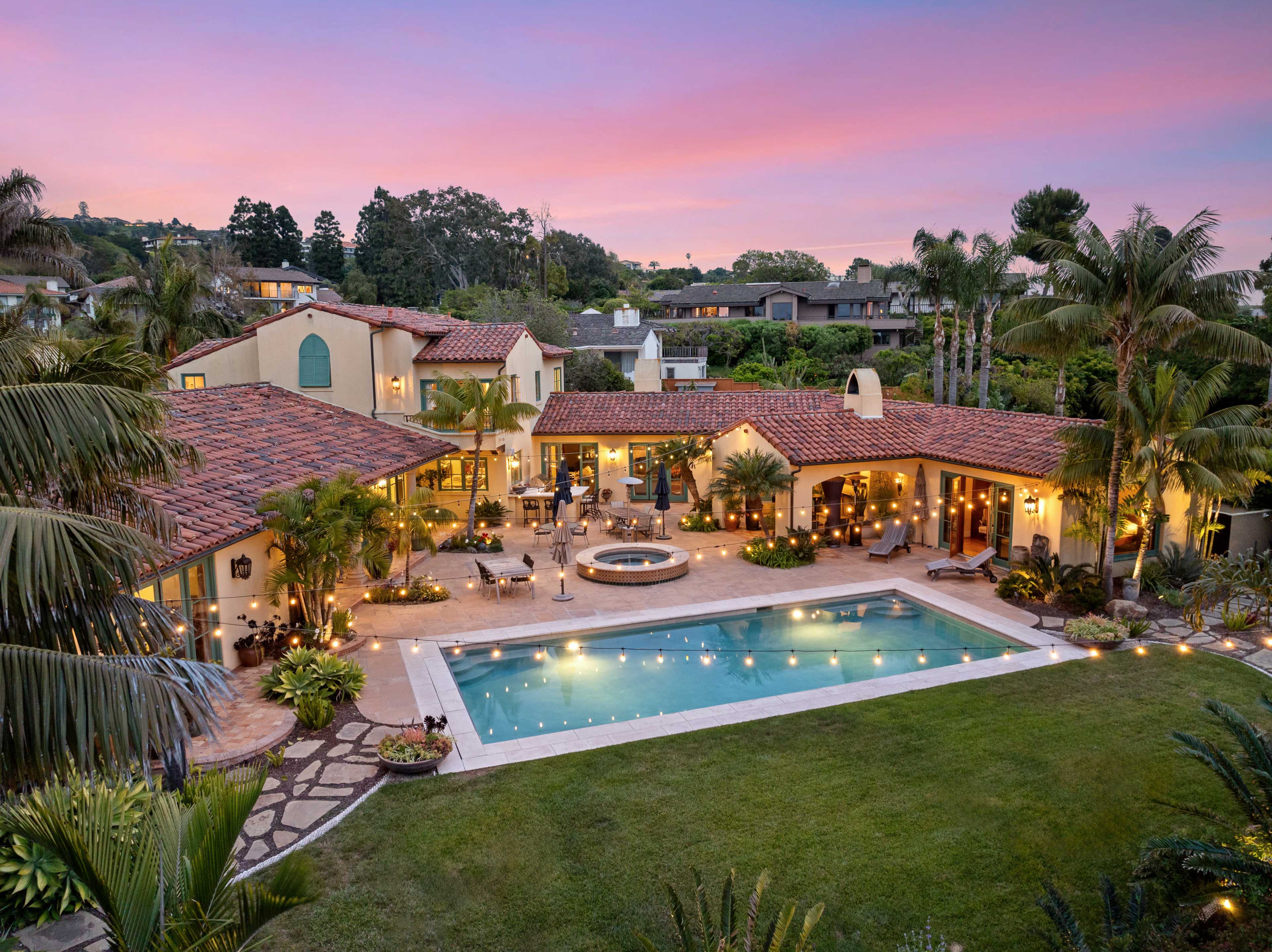 A large, well-landscaped backyard features a pool, spa, and seating area surrounded by palm trees and illuminated by string lights, with a spacious home in the background.