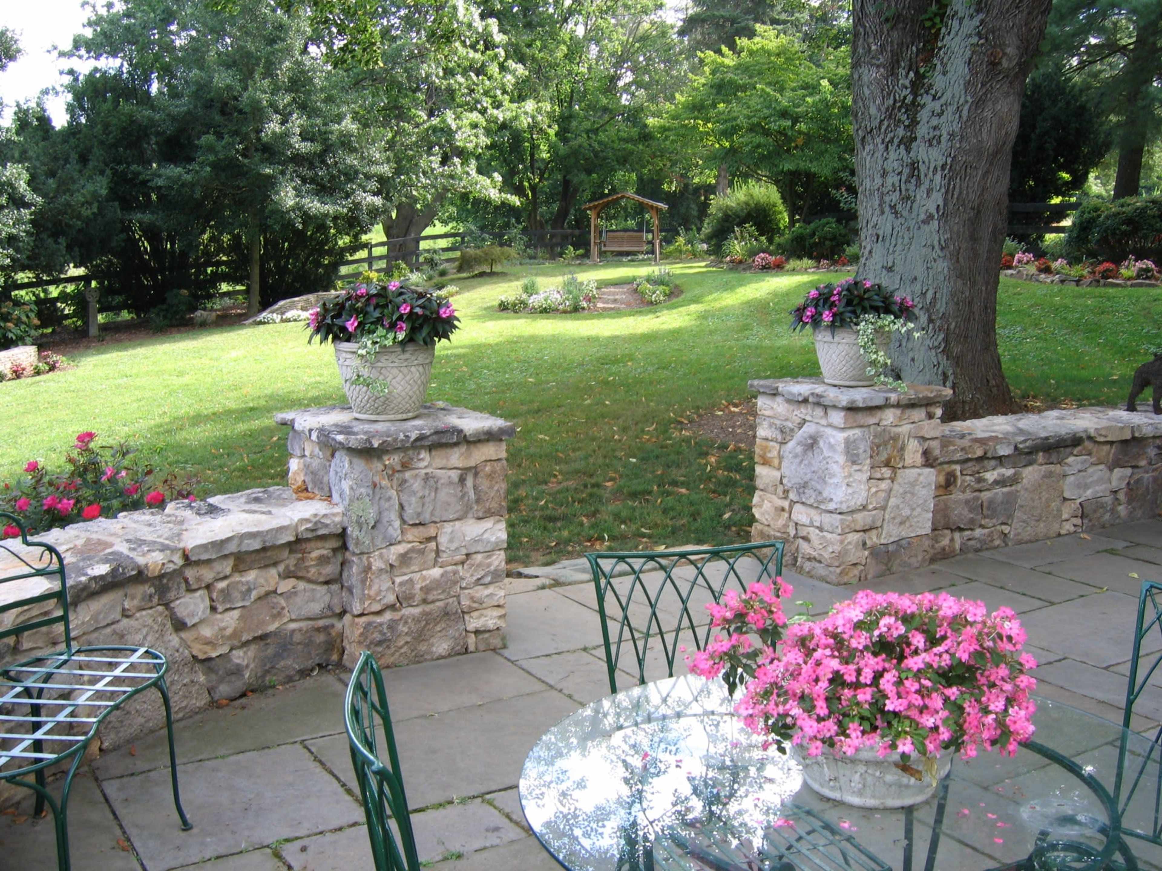 The image shows a stone patio with potted flowers and a view of a landscaped garden featuring a wooden swing in the background.