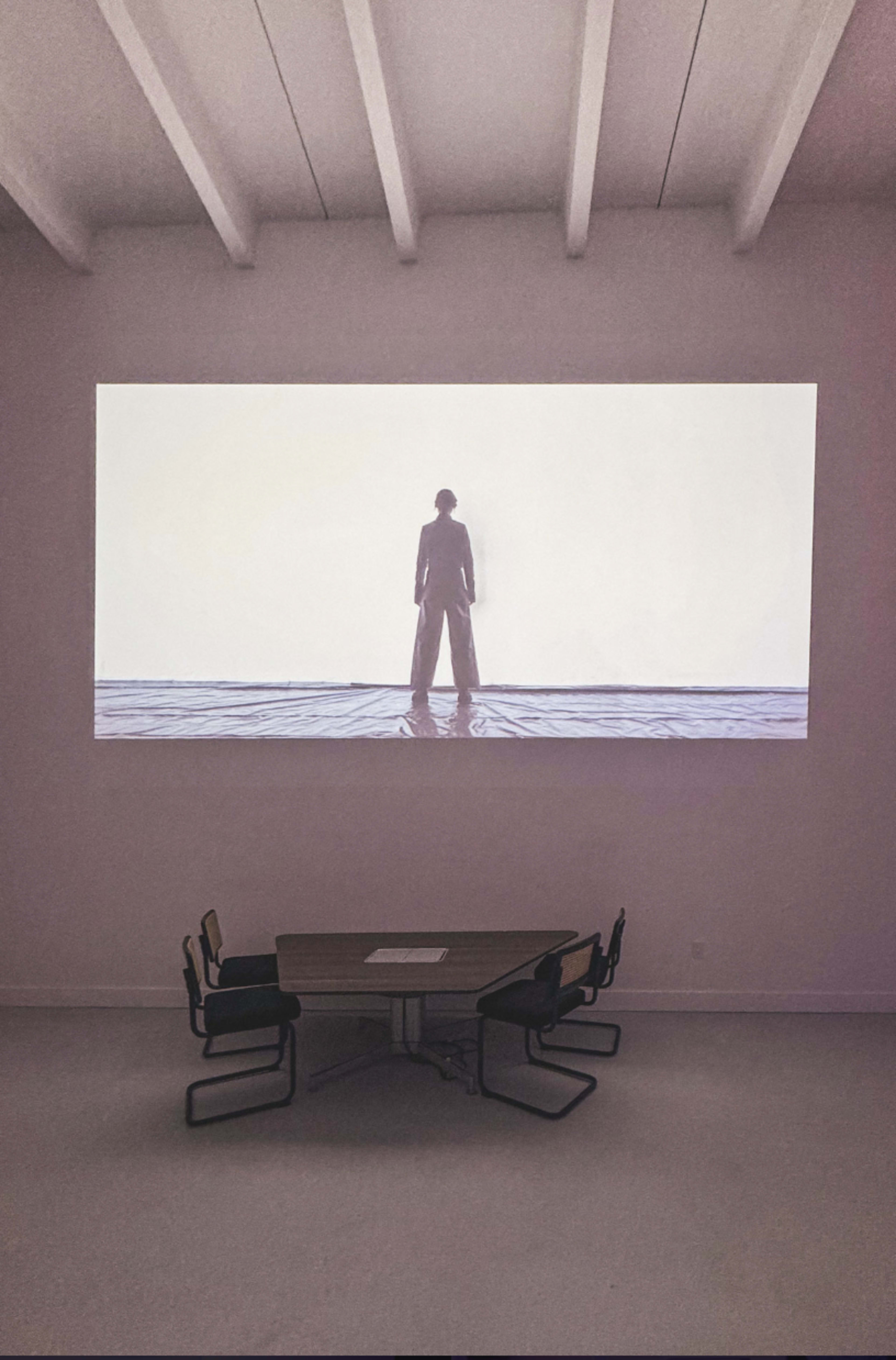 A large projection of a person standing on a beach is displayed on a wall above a table with chairs in a minimalist room.