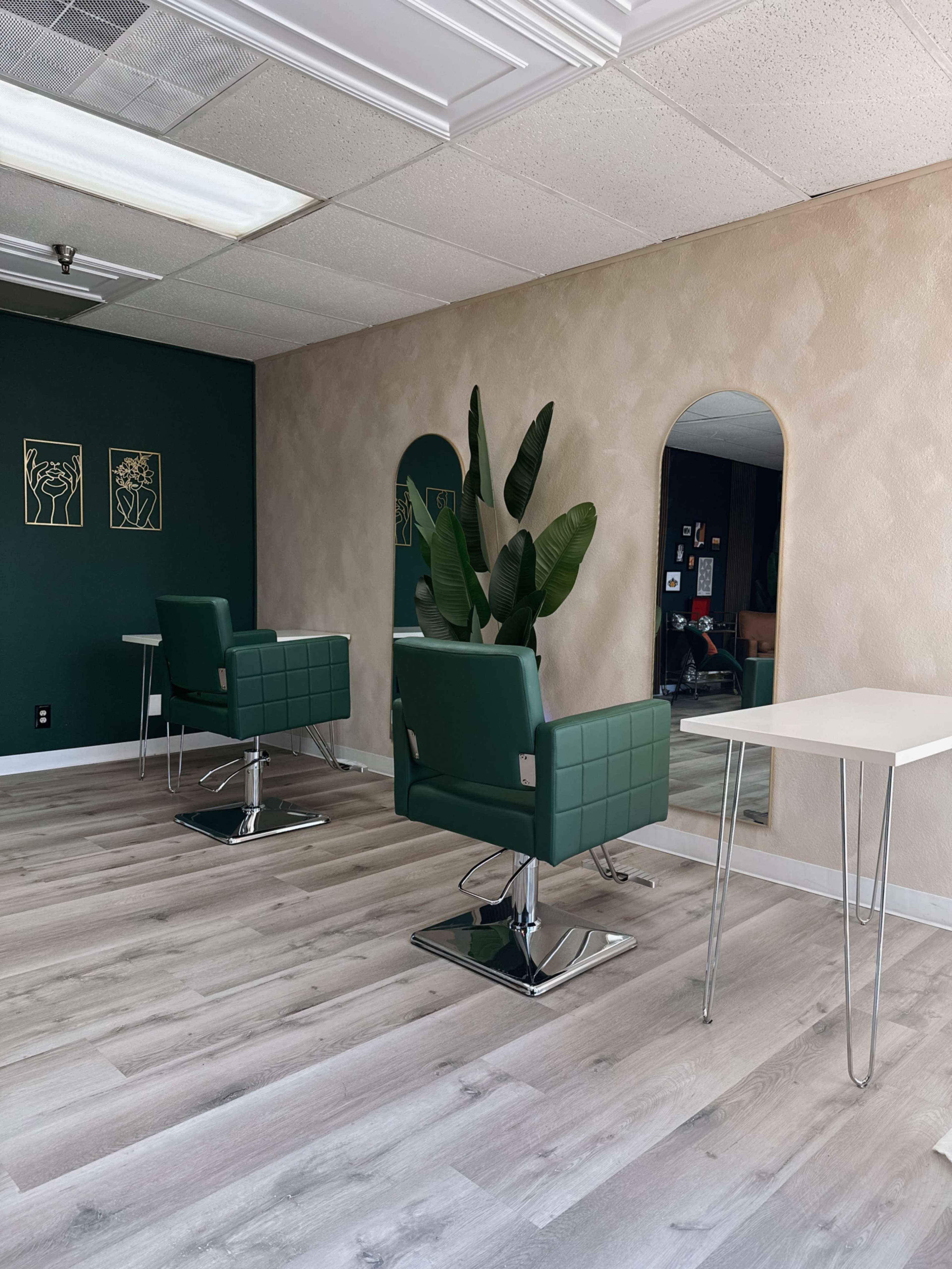The image shows a modern salon interior featuring two green styling chairs, a table, and a large plant against a textured wall.